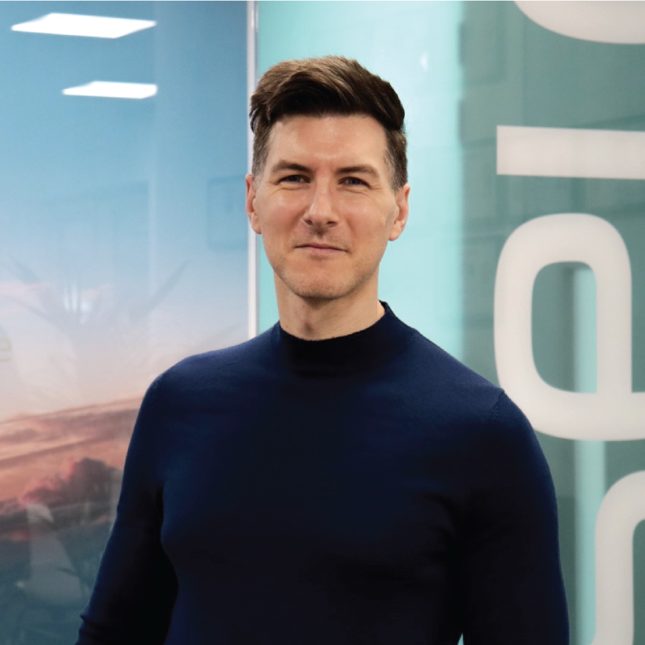 A young man with short brown hair, wearing a navy blue turtleneck sweater, standing indoors in front of a glass wall with the word 'Dell' partially visible.