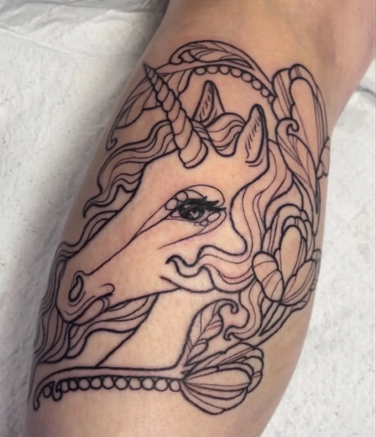 Line art tattoo of a unicorn with flowing hair and a large eye on the face, surrounded by decorative elements.