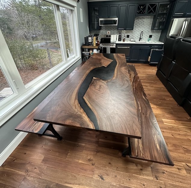A large wooden dining table with a black epoxy river running through the middle, situated in a room with hardwood floors and large windows overlooking a wooded area, with a kitchen in the background.