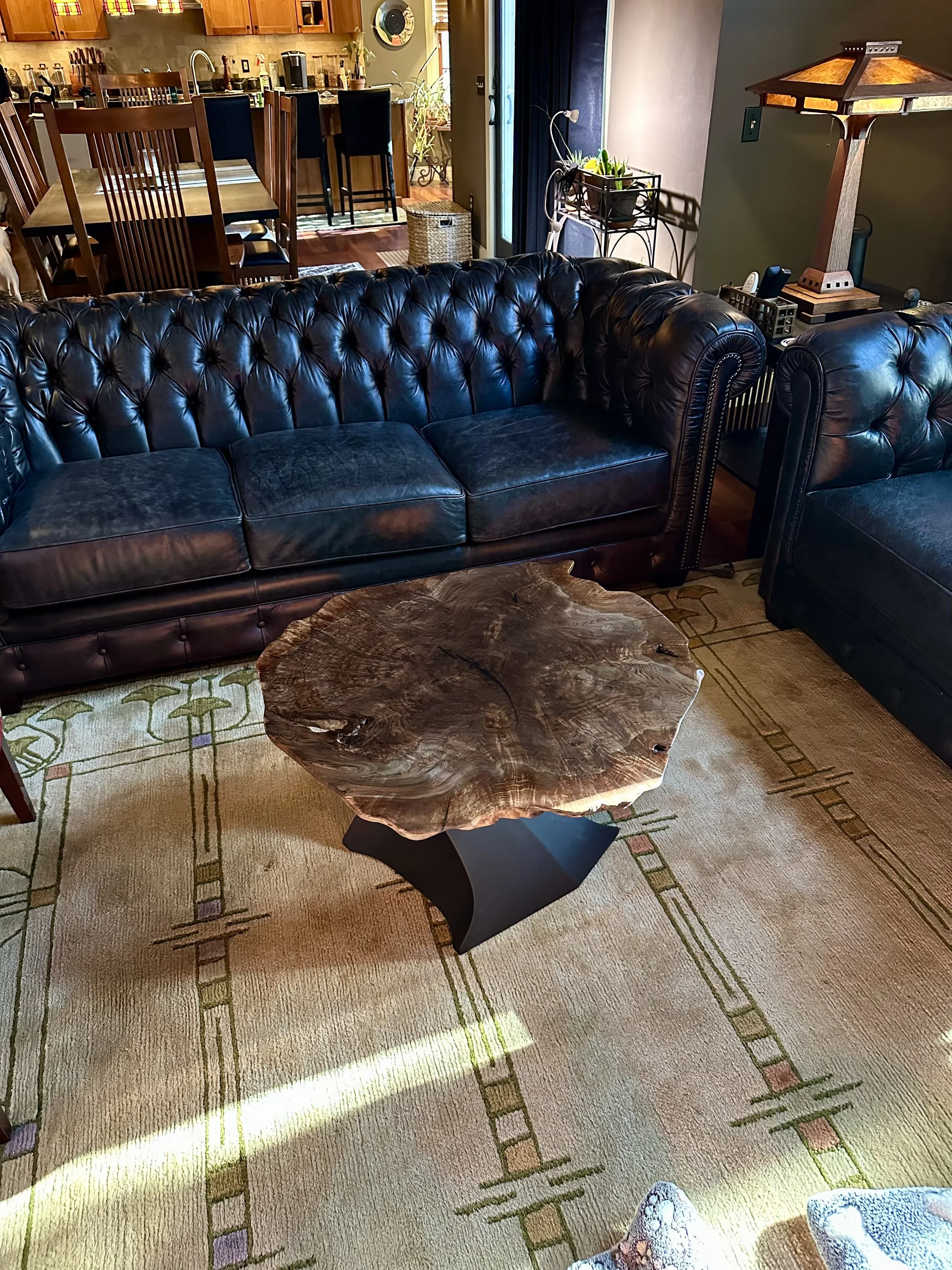 Living room with black leather sofas and a wooden slab coffee table on a patterned rug, with a Craftsman-style lamp and a view of a dining and kitchen area in the background.