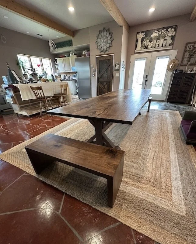 A rustic wooden dining table with a matching wooden bench in a cozy, decorated living space. The room features a patterned area rug, tile flooring, and a kitchen area in the background with open shelving and potted plants.