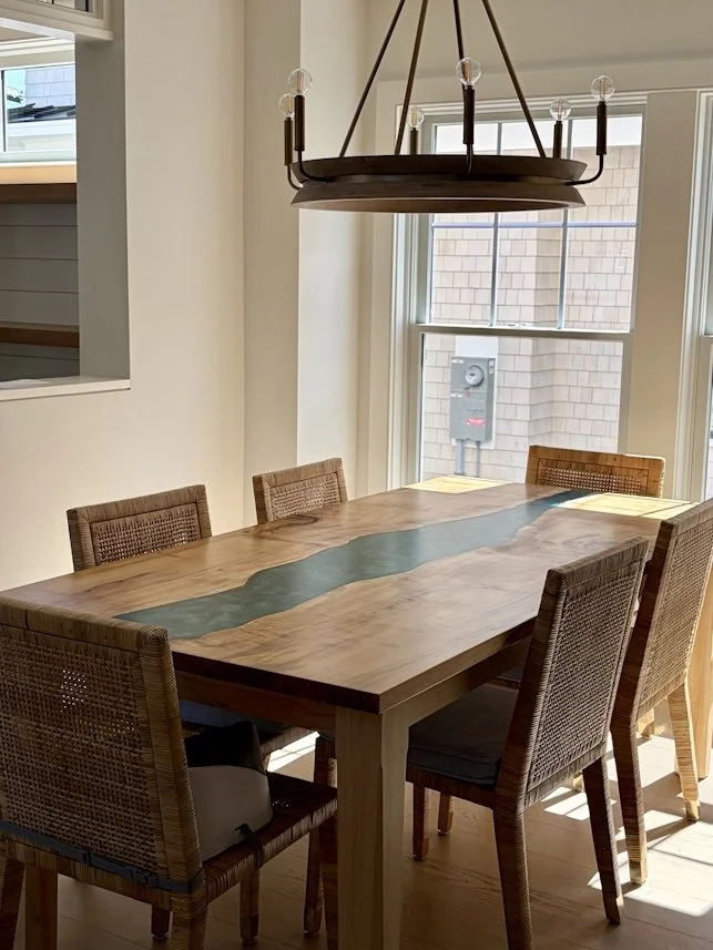 Wooden dining table with a dark green centerpiece, surrounded by six woven rattan chairs, in a room with large windows and a modern chandelier overhead.
