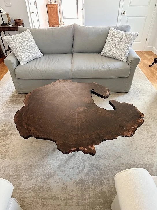 A living room with a light gray sofa, two patterned throw pillows, a rustic wooden coffee table with an irregular shape, a beige area rug, and a white wall with a door in the background.