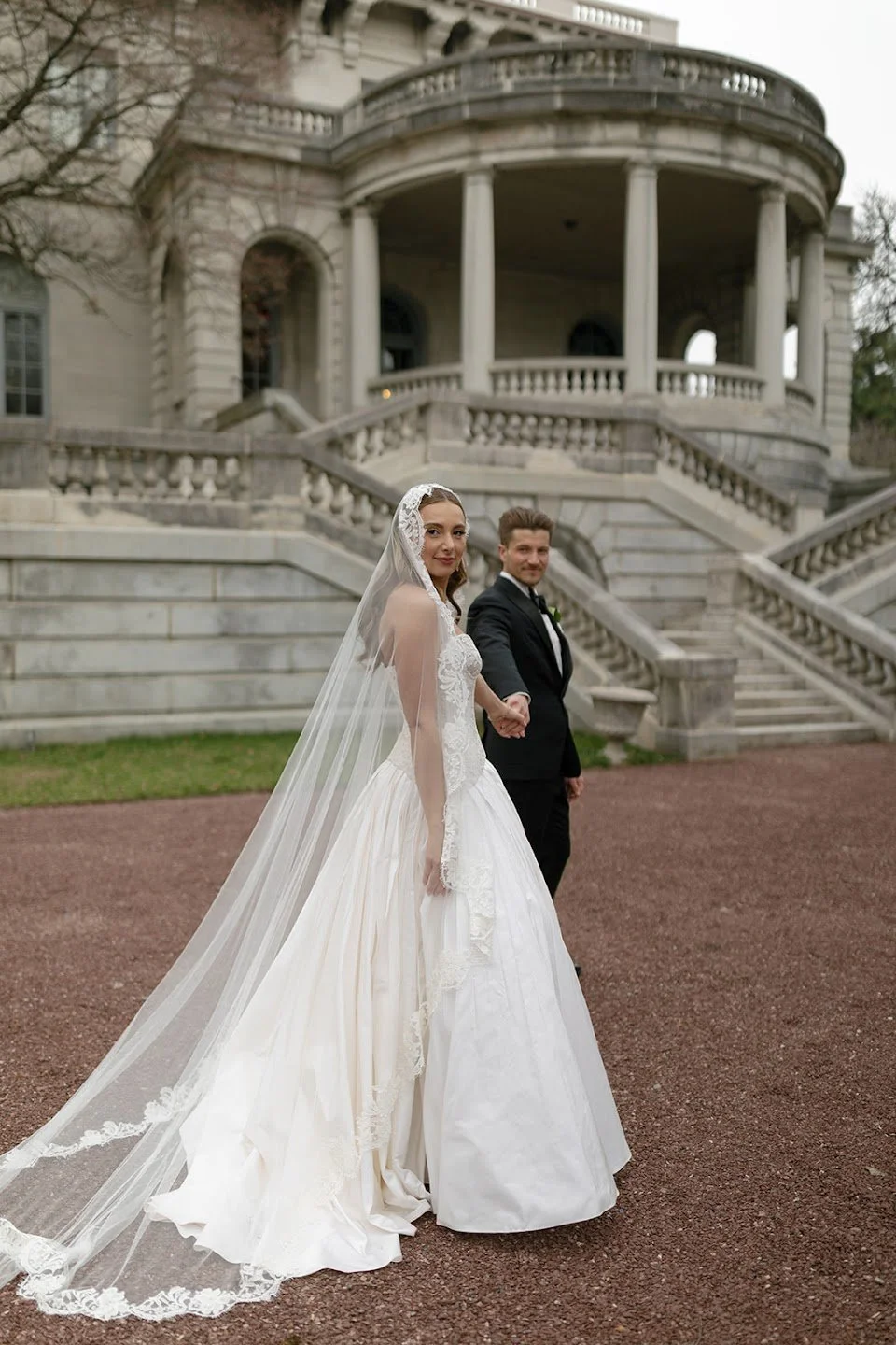 Newlywed couple walking hand in hand through a European-inspired estate in Elkins Park, PA. Documentary-style wedding photography featuring authentic moments, elegant architecture, timeless wedding fashion, and natural movement.