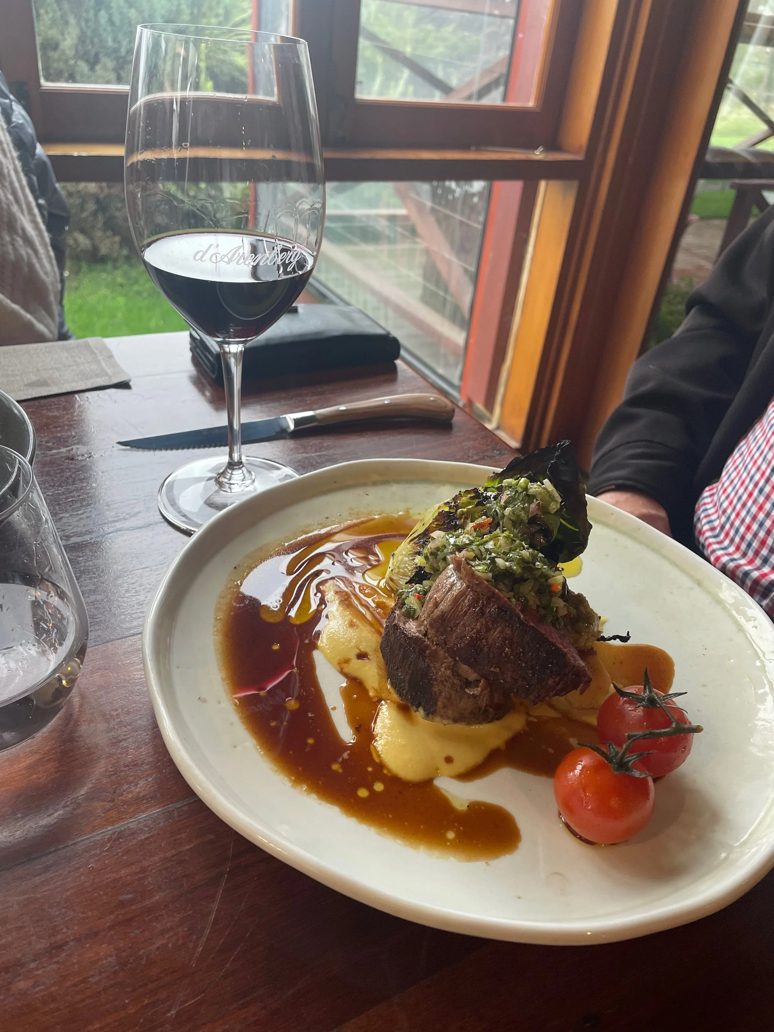 A plate of food with a beef steak topped with pico de gallo, served with mashed potatoes and a cherry tomato on the side. In the background, there is a glass of red wine on a wooden table near a window.