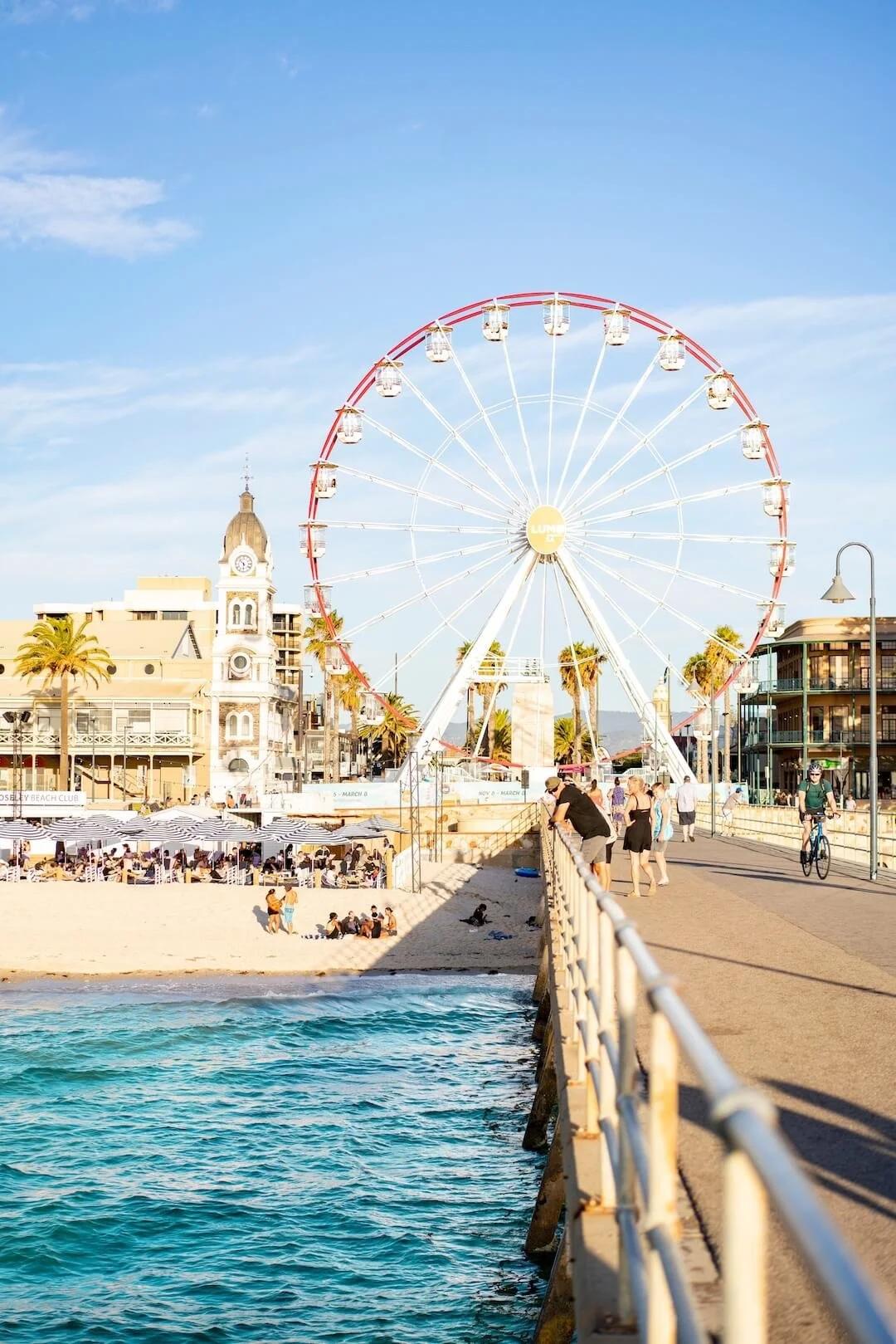 A seaside boardwalk with a large Ferris wheel, a church, palm trees, and beachgoers relaxing on the sand and walking along the pier during the day.