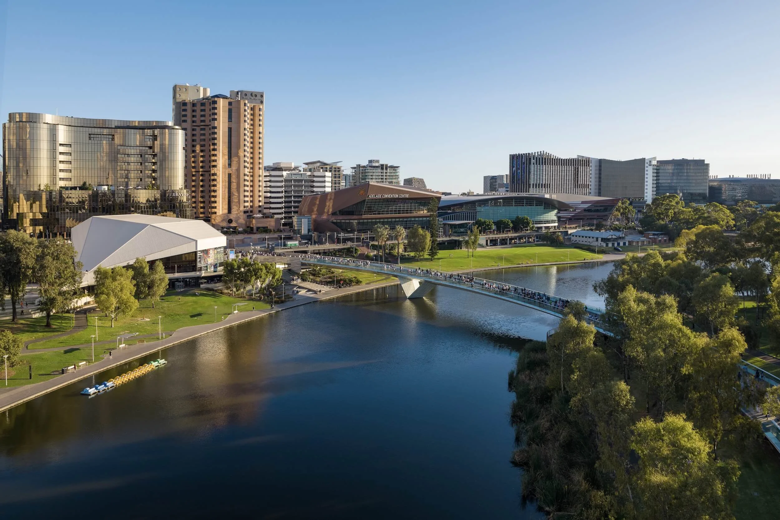 A cityscape featuring modern high-rise buildings near a body of water with a park and a bridge in the foreground, under a clear blue sky.