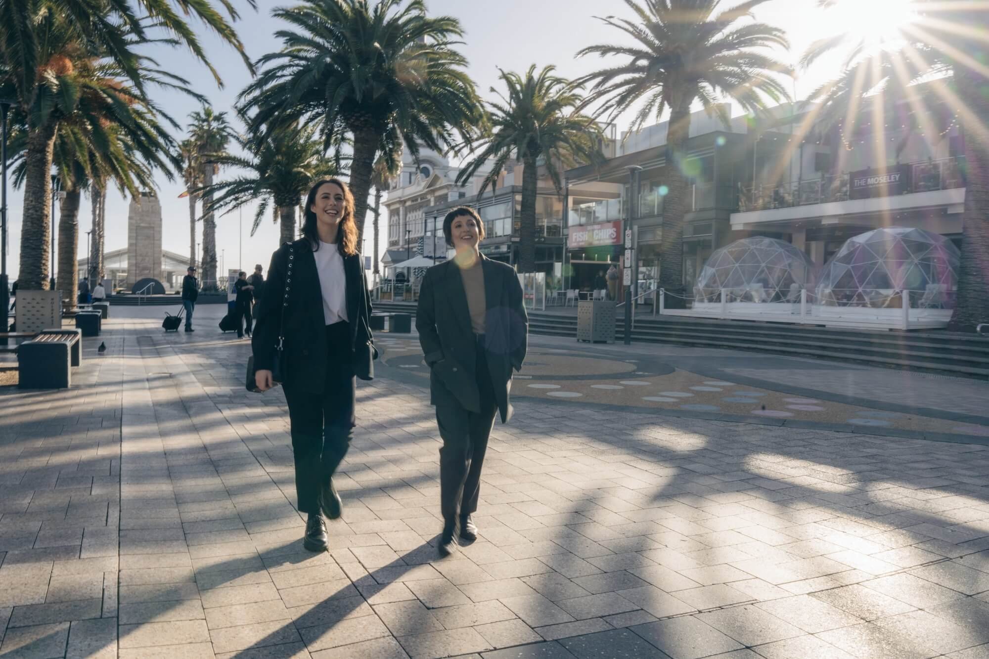 Two women walking on a sunny promenade lined with tall palm trees, with retail shops and outdoor seating in the background.