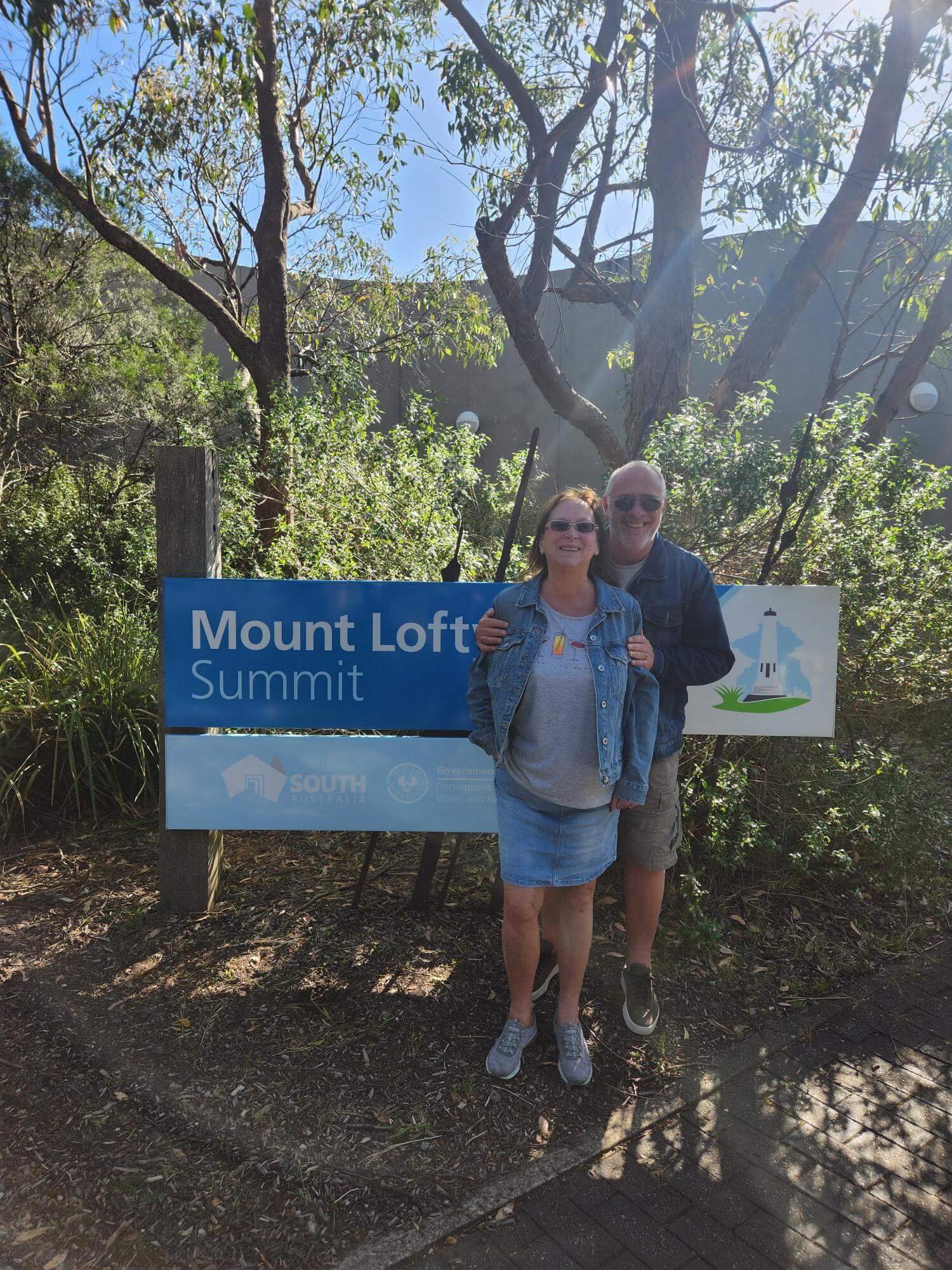 A couple standing smiling in front of a sign that reads 'Mount Lofty Summit' with a lighthouse illustration. The background features trees, a clear blue sky, and sunlight filtering through the branches.