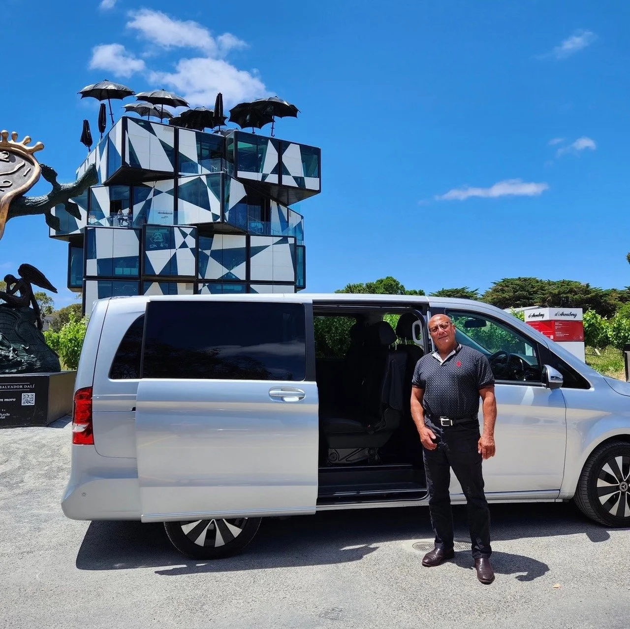 A man standing outside of a silver van with its sliding door open, in front of a modern, geometric building with an umbrella sculpture on top, under a partly cloudy blue sky.