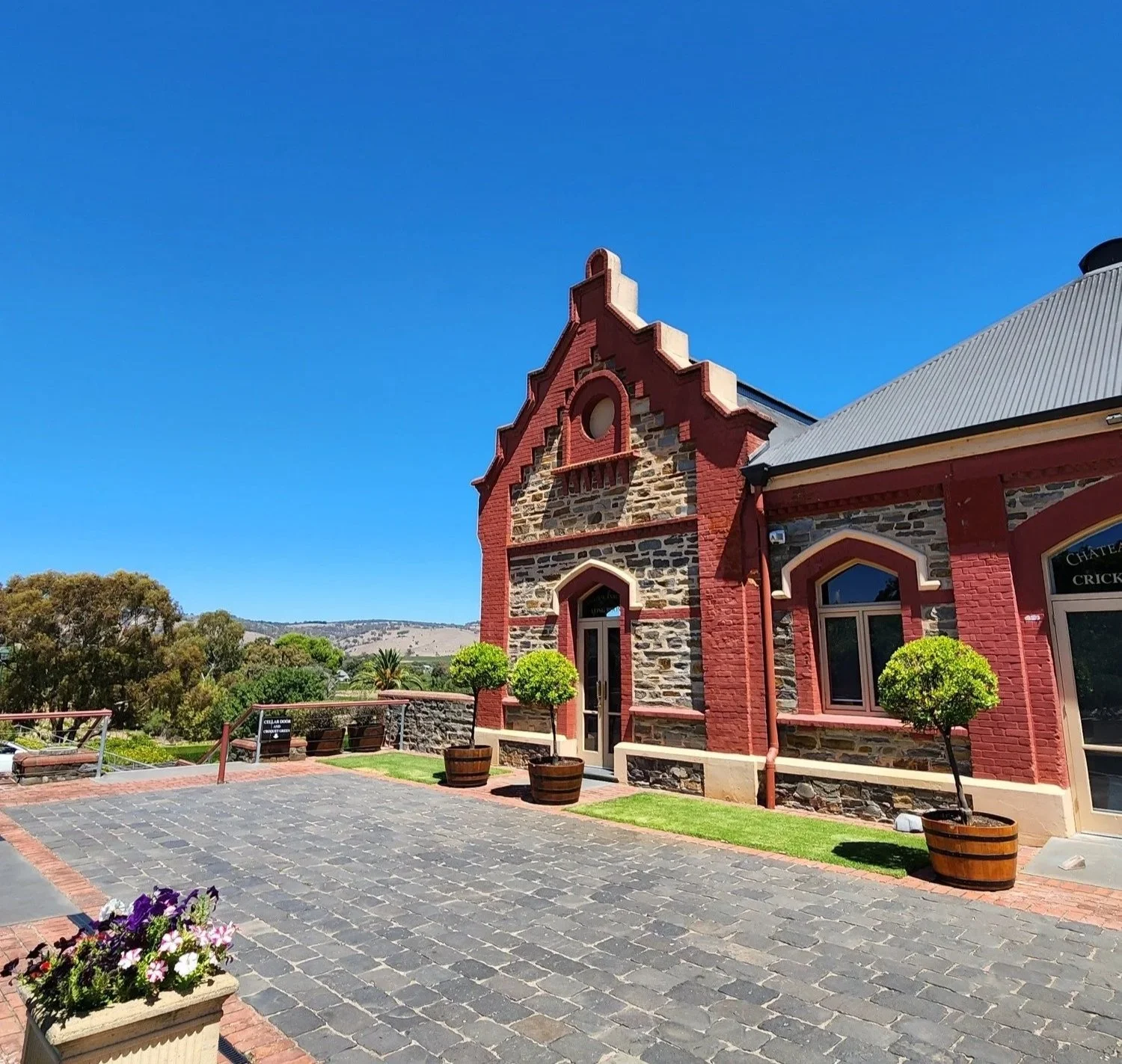 A historic red brick building with stone accents and arched windows, situated in a landscaped area with potted trees and flowering plants under a clear blue sky.