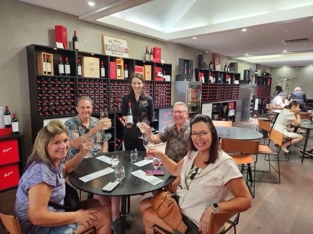 Five people sitting around a table and toasting with wine glasses in a wine cellar or tasting room. The background shows wine bottles on shelves and other patrons dining.
