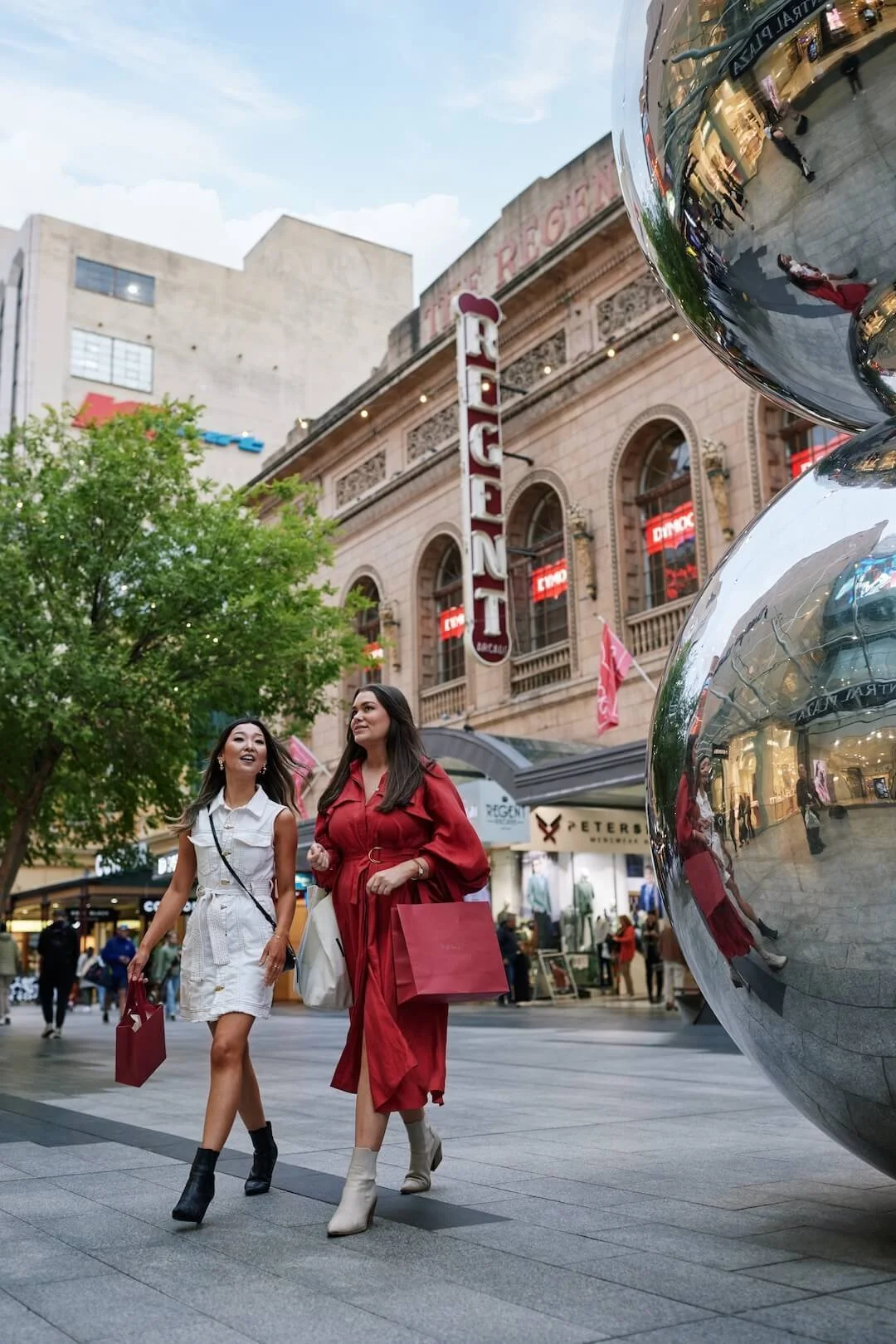 Two women with shopping bags walking in a city shopping district, with a theater sign and store windows in the background.