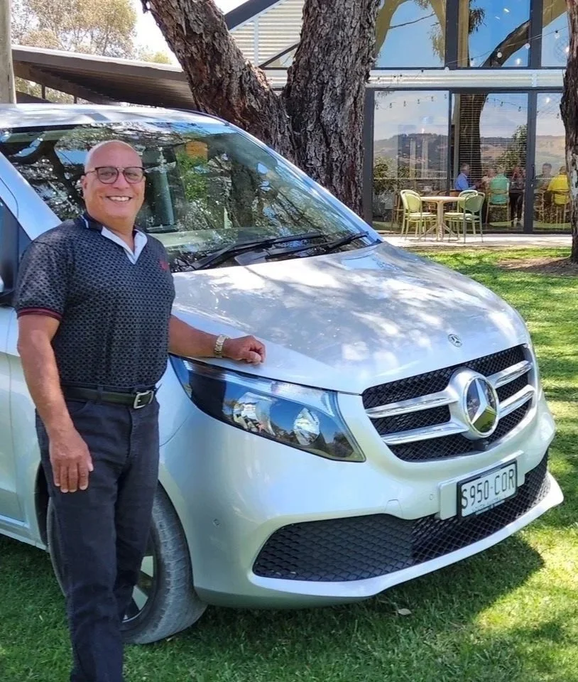 Man standing beside a silver Mercedes-Benz van with a license plate reading 'S950-COR,' outdoors with trees and a building in the background, some people sitting at a table inside visible through glass.