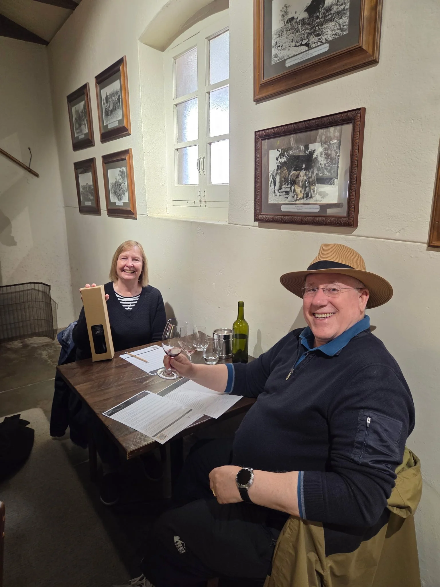 A man and a woman sitting at a dining table in a restaurant, smiling and enjoying wine. The man is wearing a wide-brimmed hat and glasses, holding a glass of red wine. The woman has shoulder-length blond hair, is smiling, and appears to be holding a menu or a brochure. The table has a bottle of green wine, several glasses, and papers. The background features a cream-colored wall with framed black-and-white photographs and a window with frosted glass.
