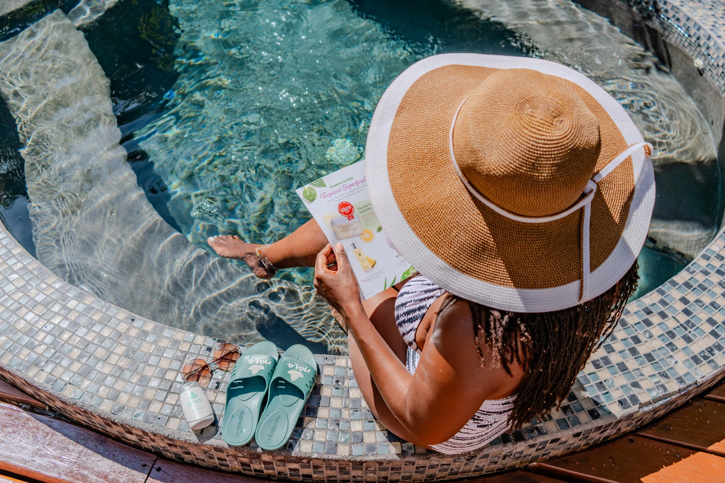 Overhead view of a guest with a sunhat reading at the Naia spa pool, featuring Naia branded sunscreen, lotion, and slides by the water's edge