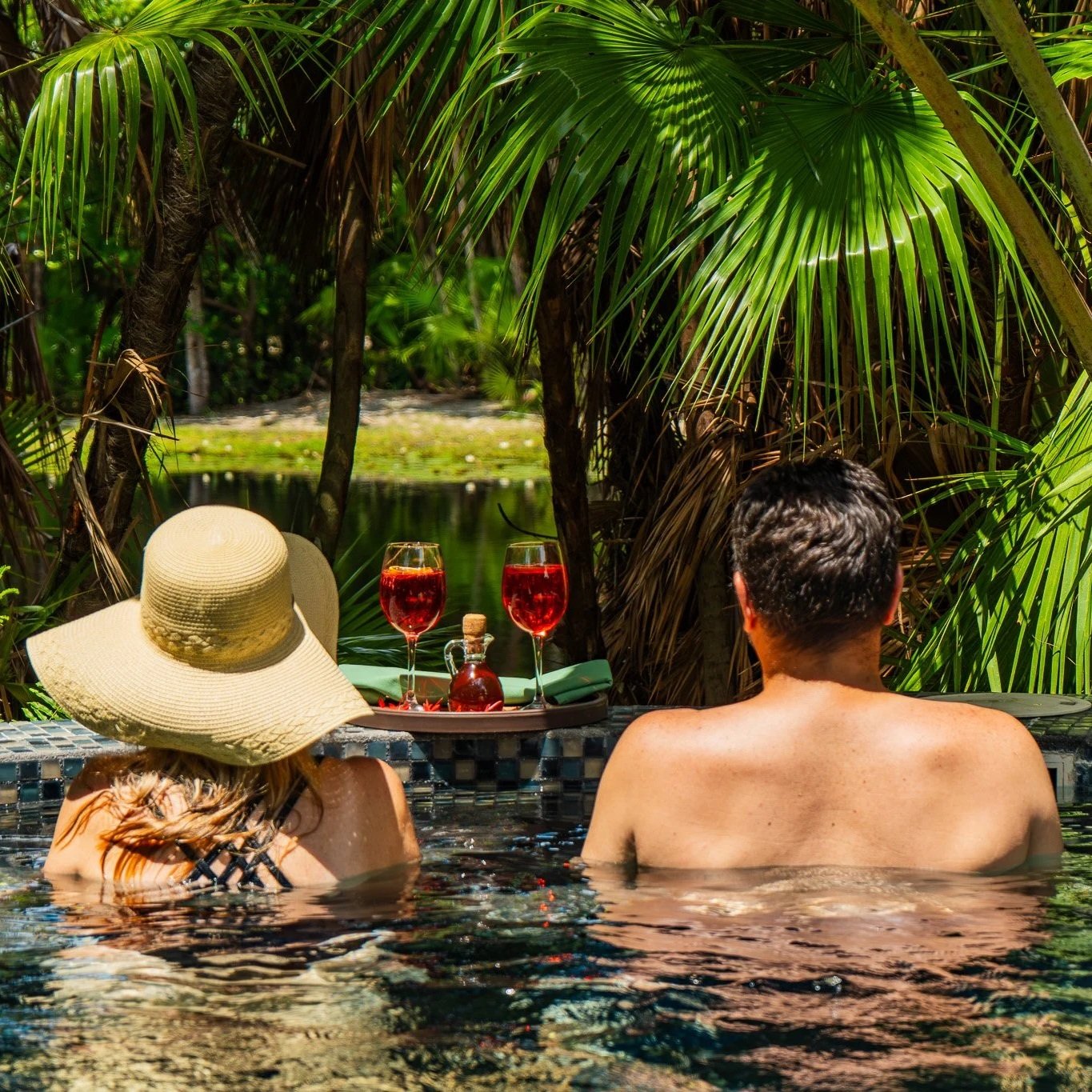 A tropical beach scene with clear turquoise water, several boats docked near the shore, and people swimming and relaxing. There are palm trees, beach huts, and people walking on the sandy beach.