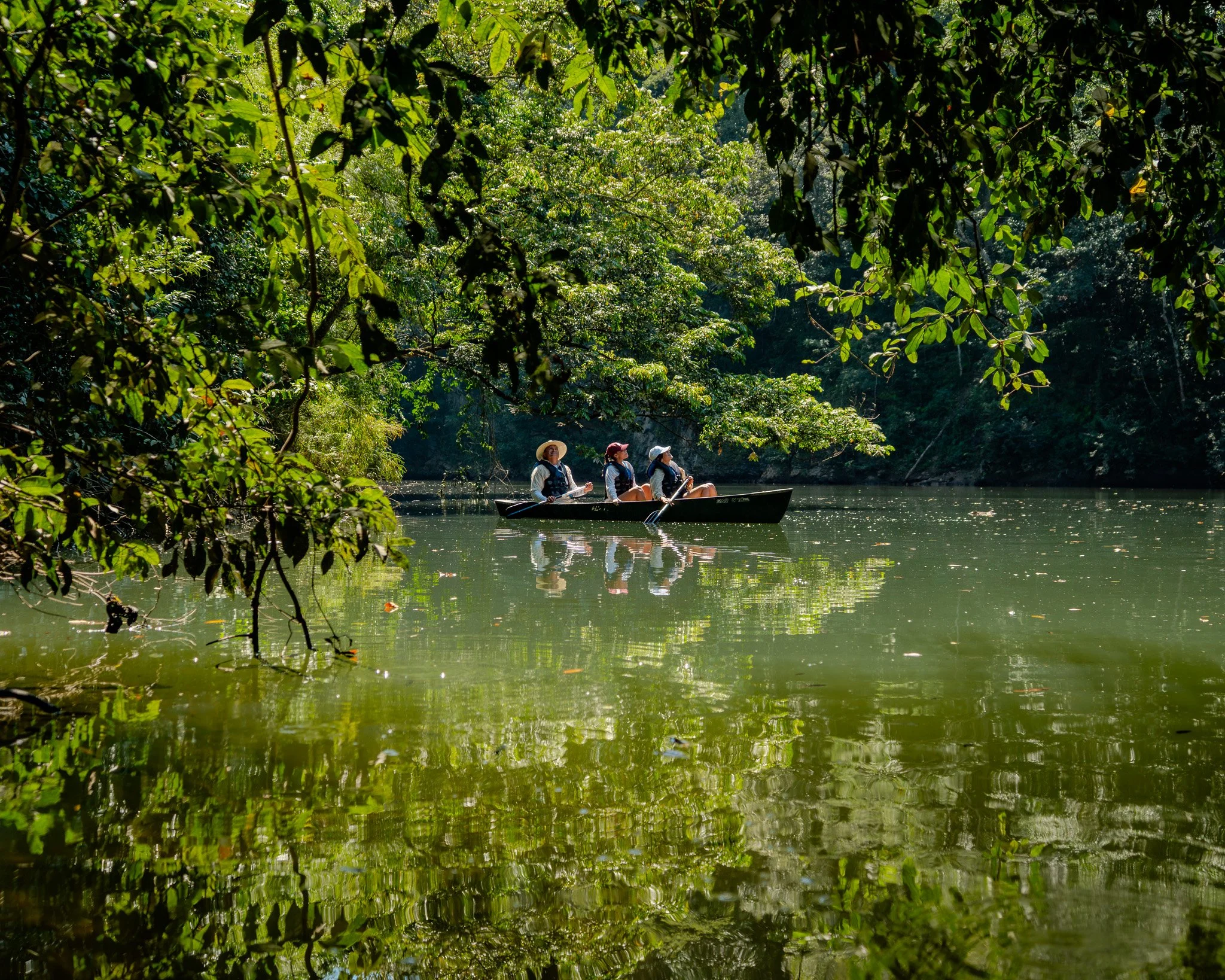 Two female guests on a guided Macaw river canoe tour with a professional guide from San Ignacio Resort Hotel, framed by lush green trees on the river bank.
