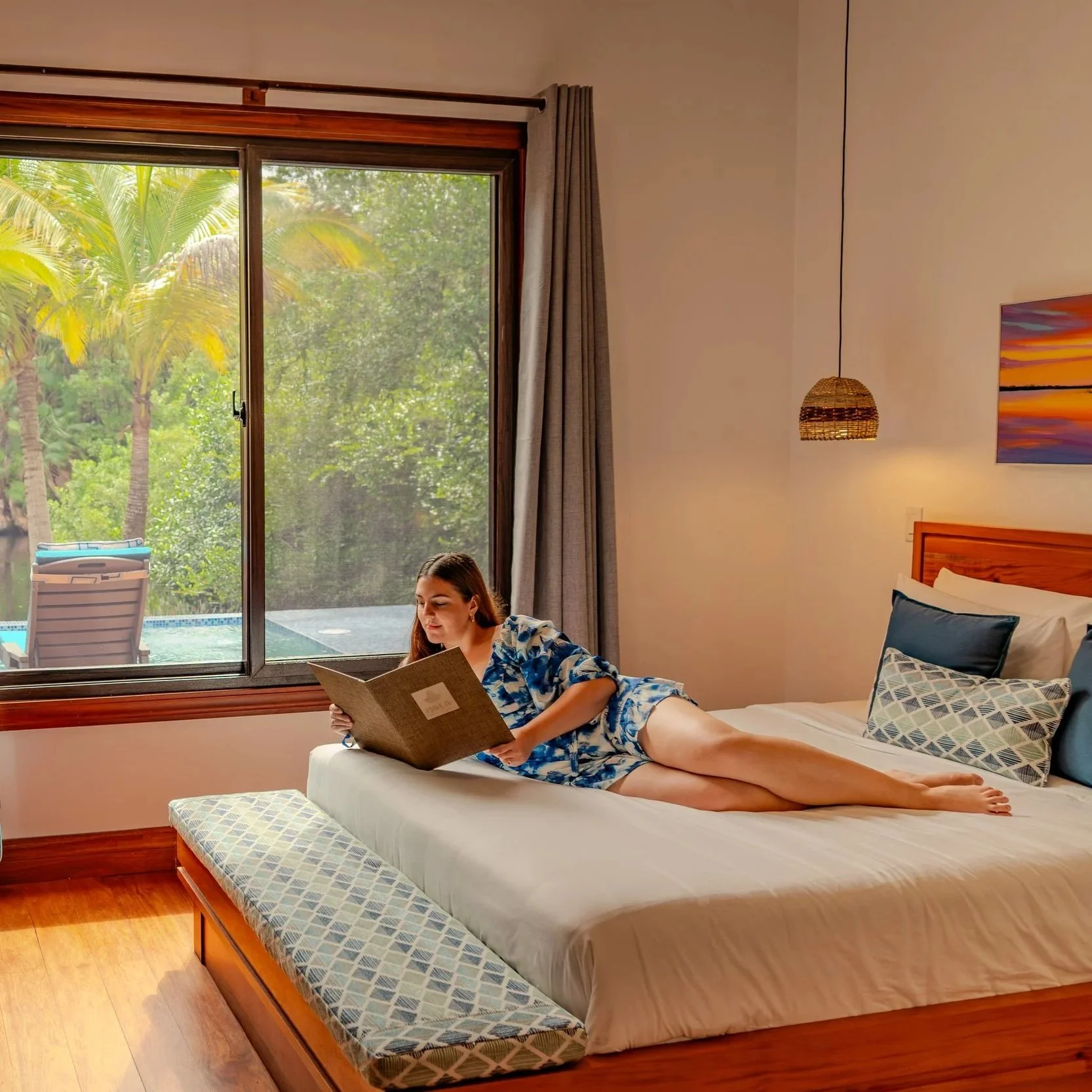 A guest relaxes on the bed of a new luxury Spa Villa at Naia Resort, looking at a spa menu with a private plunge pool and the scenic lagoon creek in the background.