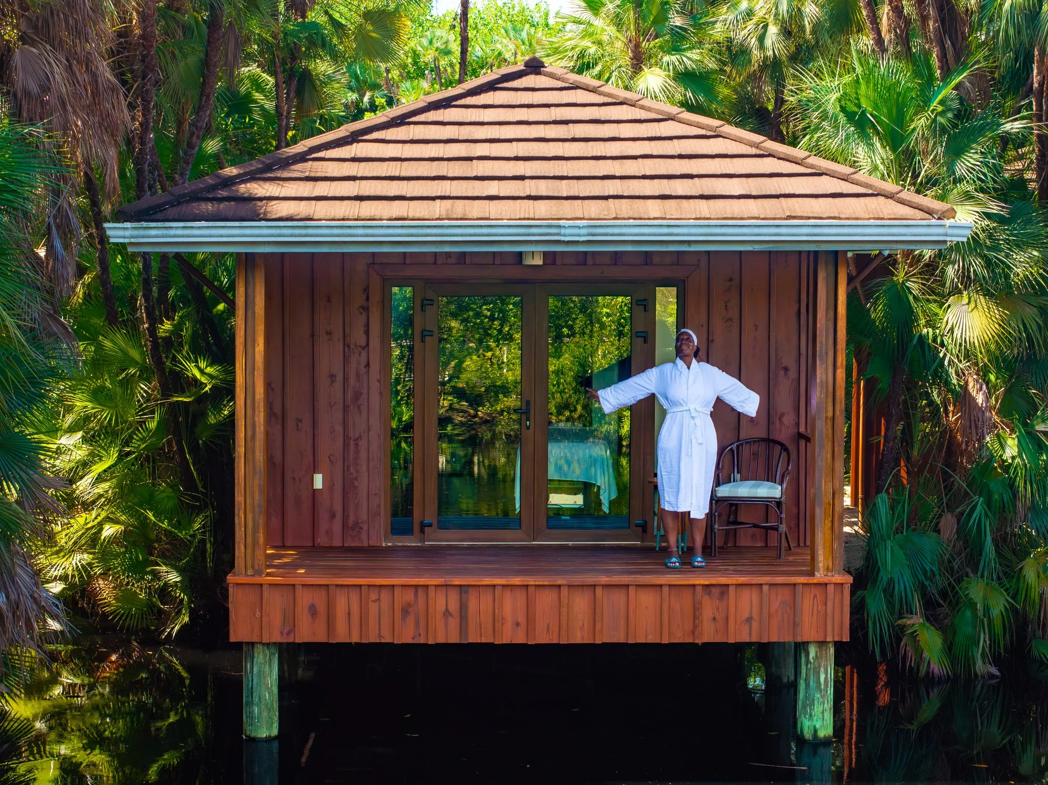 A shirtless man in black shorts standing on a rock in front of a waterfall in a lush green setting.