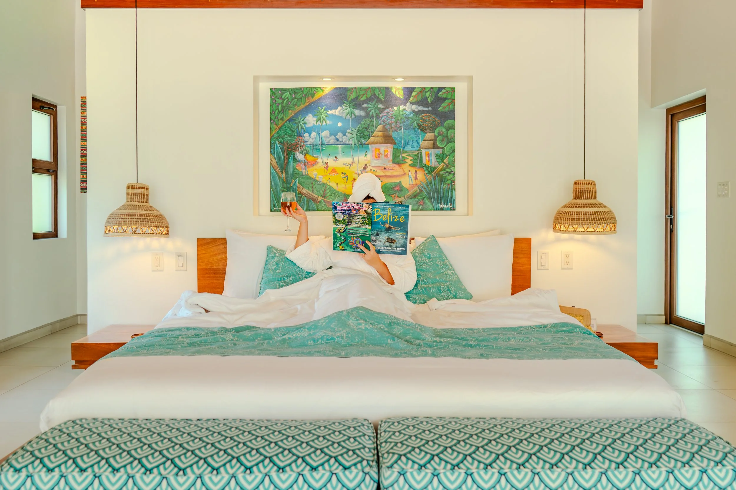 A guest relaxing in a beachfront villa at Naia Resort and Spa with a glass of wine and a Belize magazine, featuring a front-facing view of the bed and the Caribbean Sea.