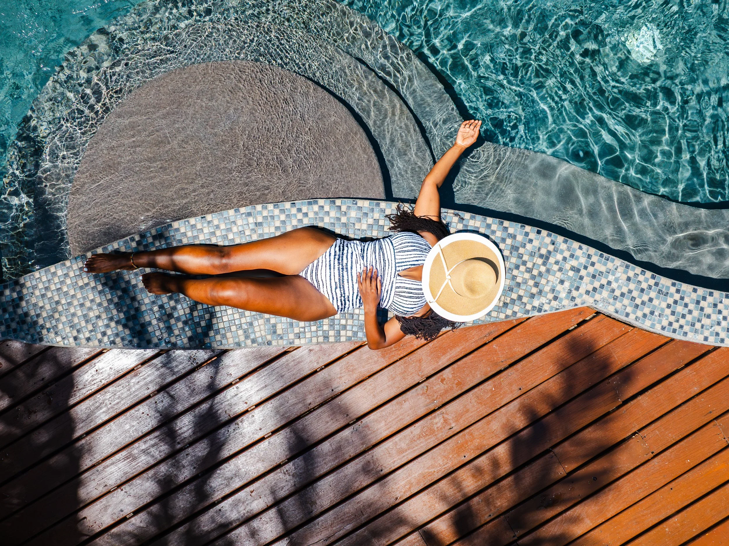 A drone view from above of a guest relaxing on the edge of the Naia Resort pool, with a sunhat on her face and one hand trailing in the turquoise water.