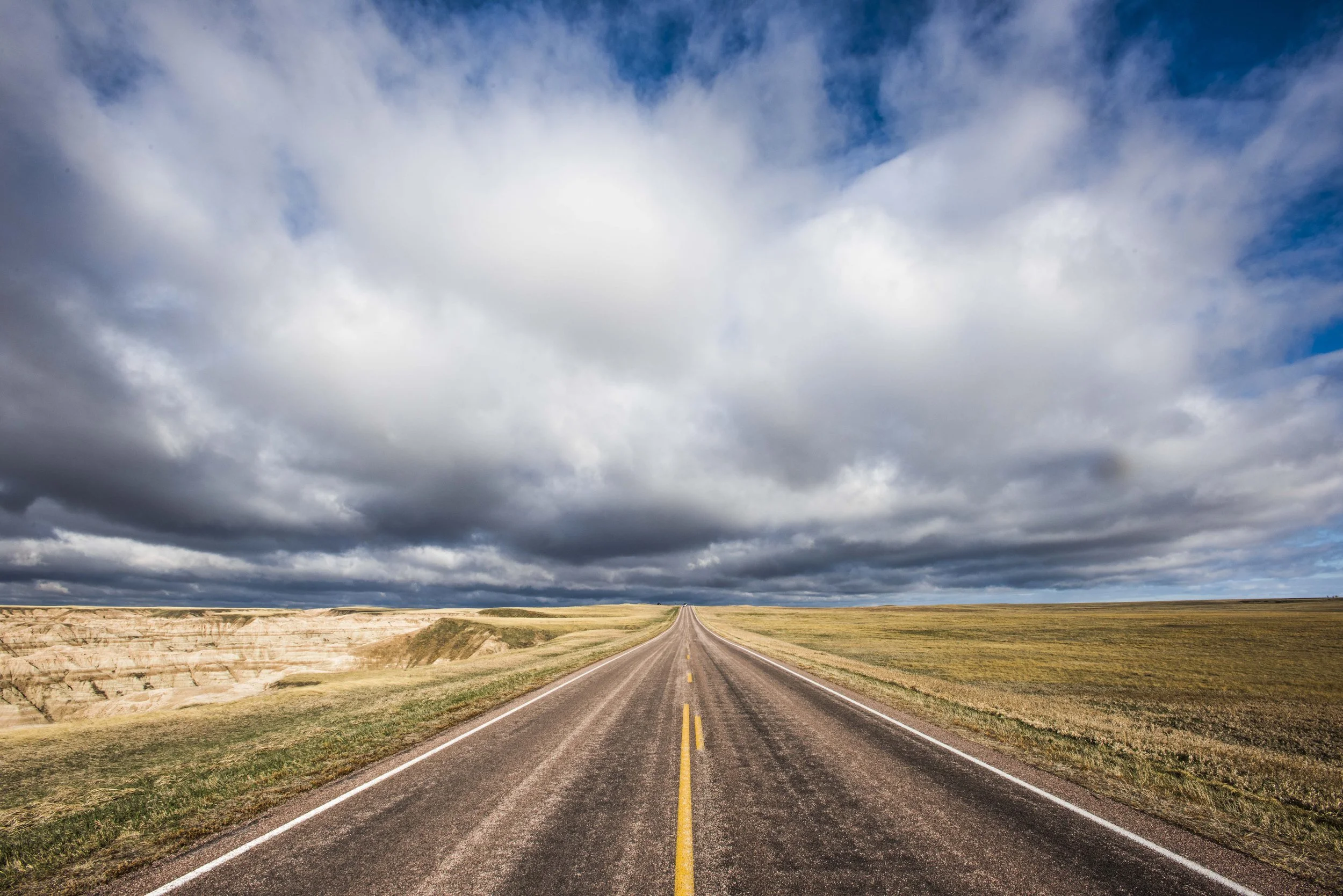 Open straight road with cloudy sky and vast grassy landscape.