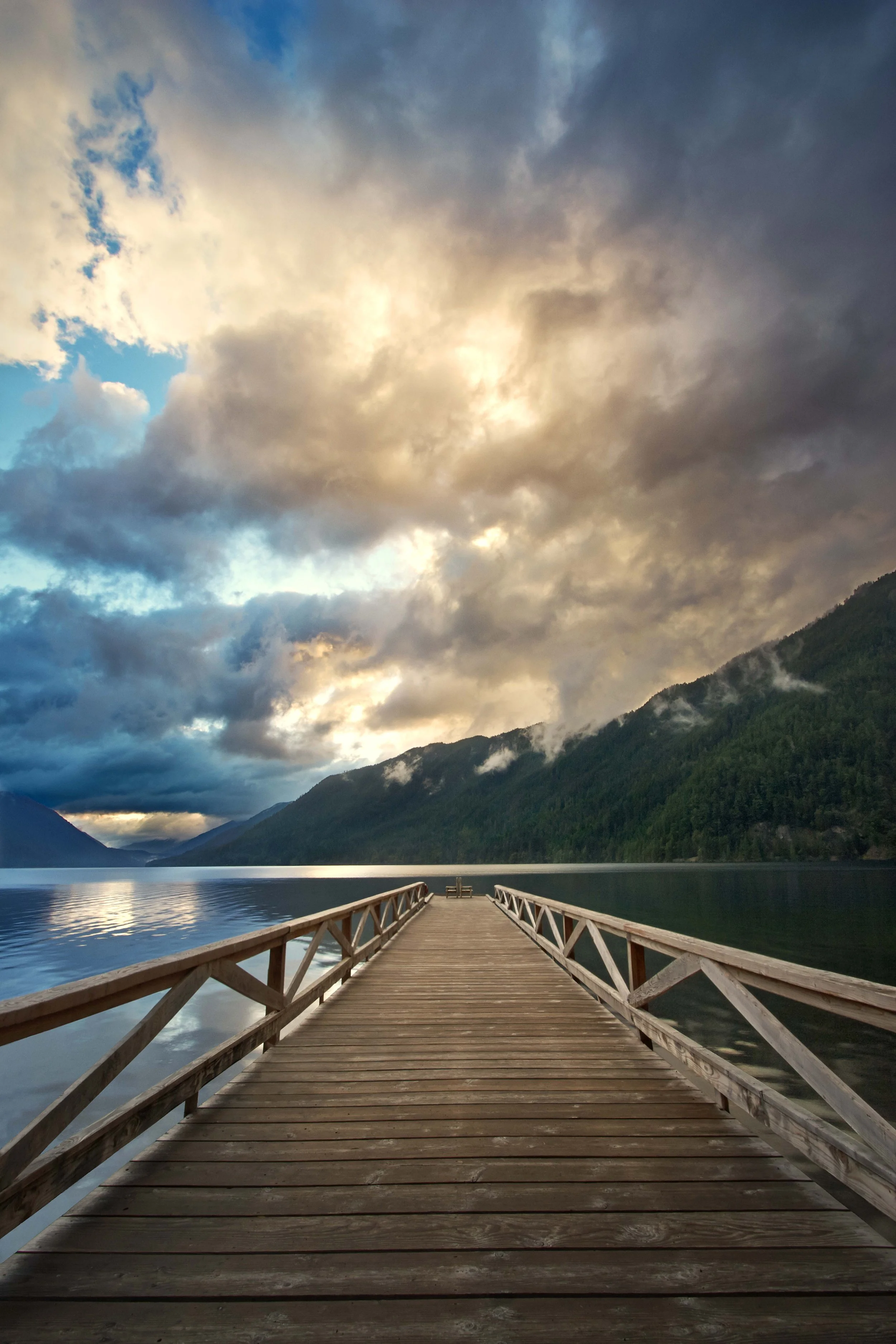 Wooden dock leading into a calm lake, surrounded by forested mountains and dramatic, cloudy sky at sunrise or sunset.
