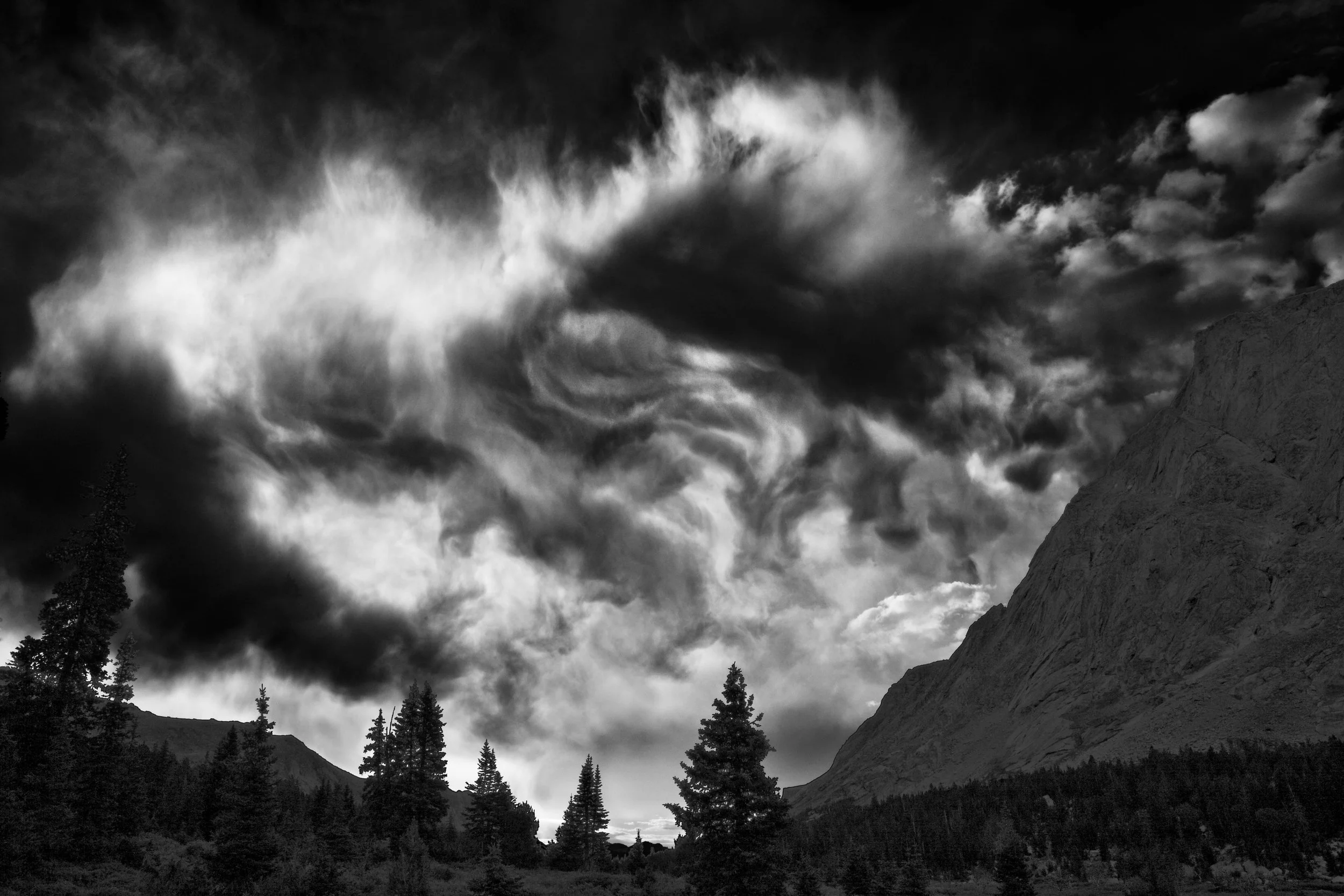 Dramatic black and white landscape with swirling clouds above a mountain and forest.