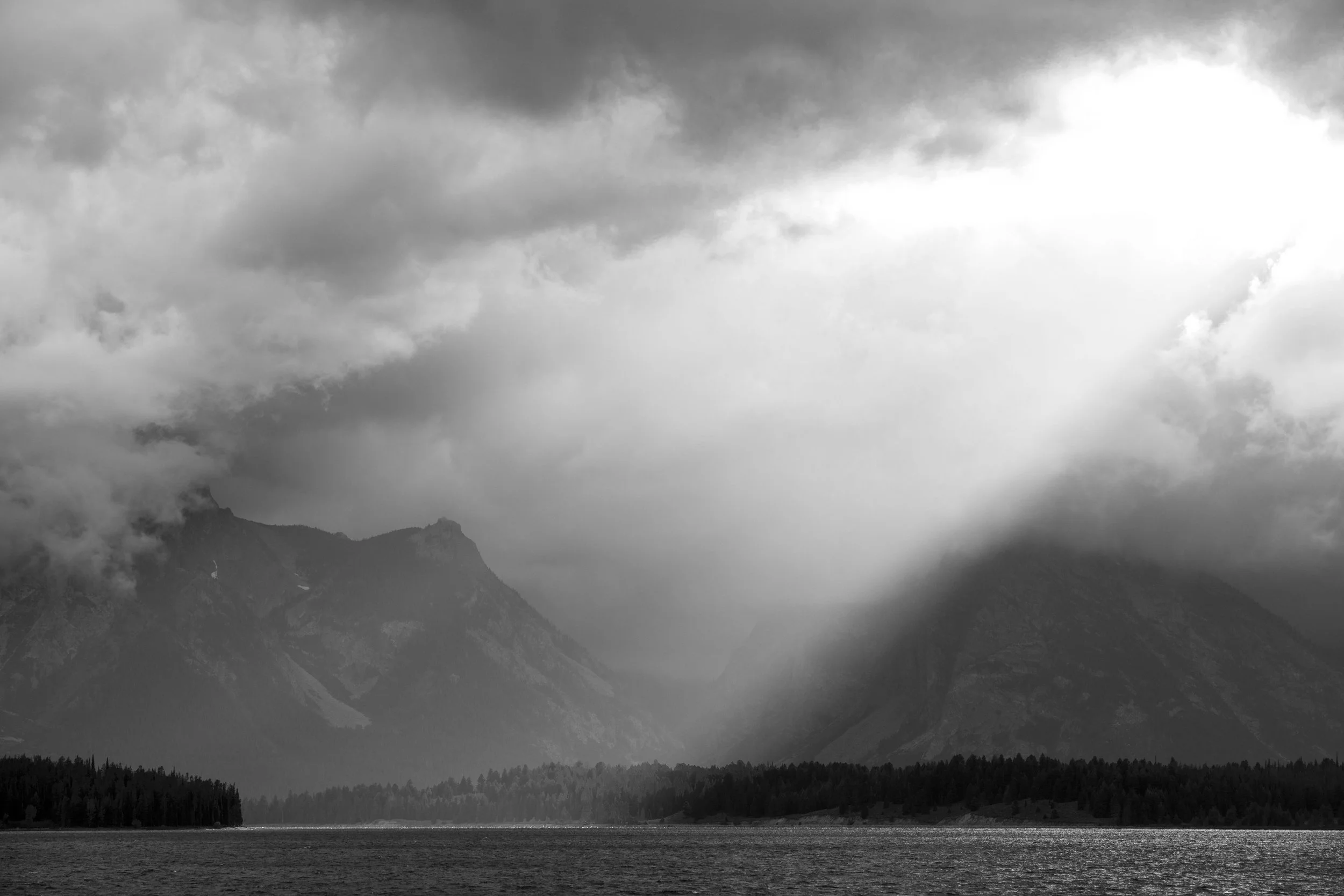 Black and white landscape of mountains shrouded in clouds with sunlight breaking through, and a lake in the foreground.