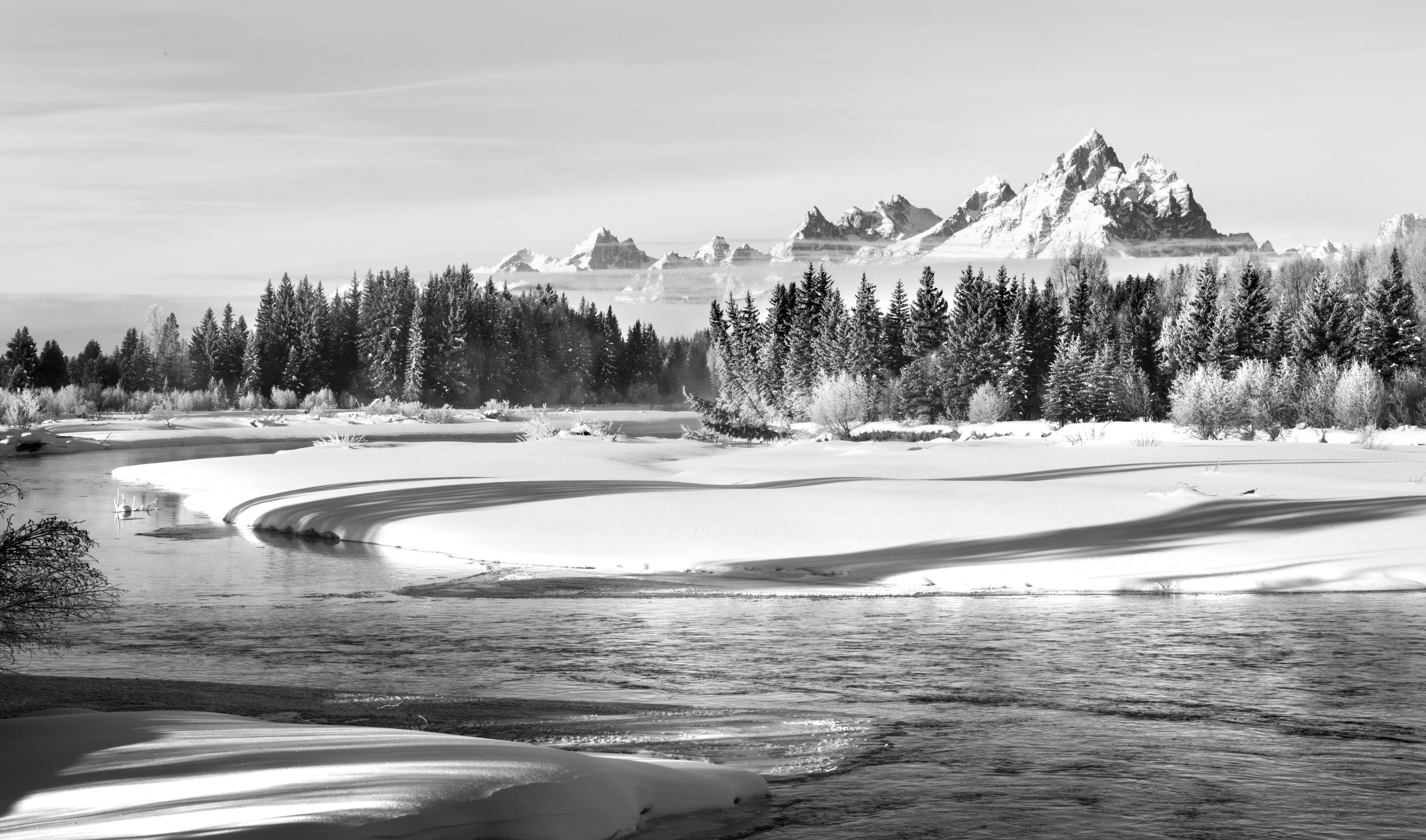 Snow-covered landscape with a river, pine trees, and distant mountains.