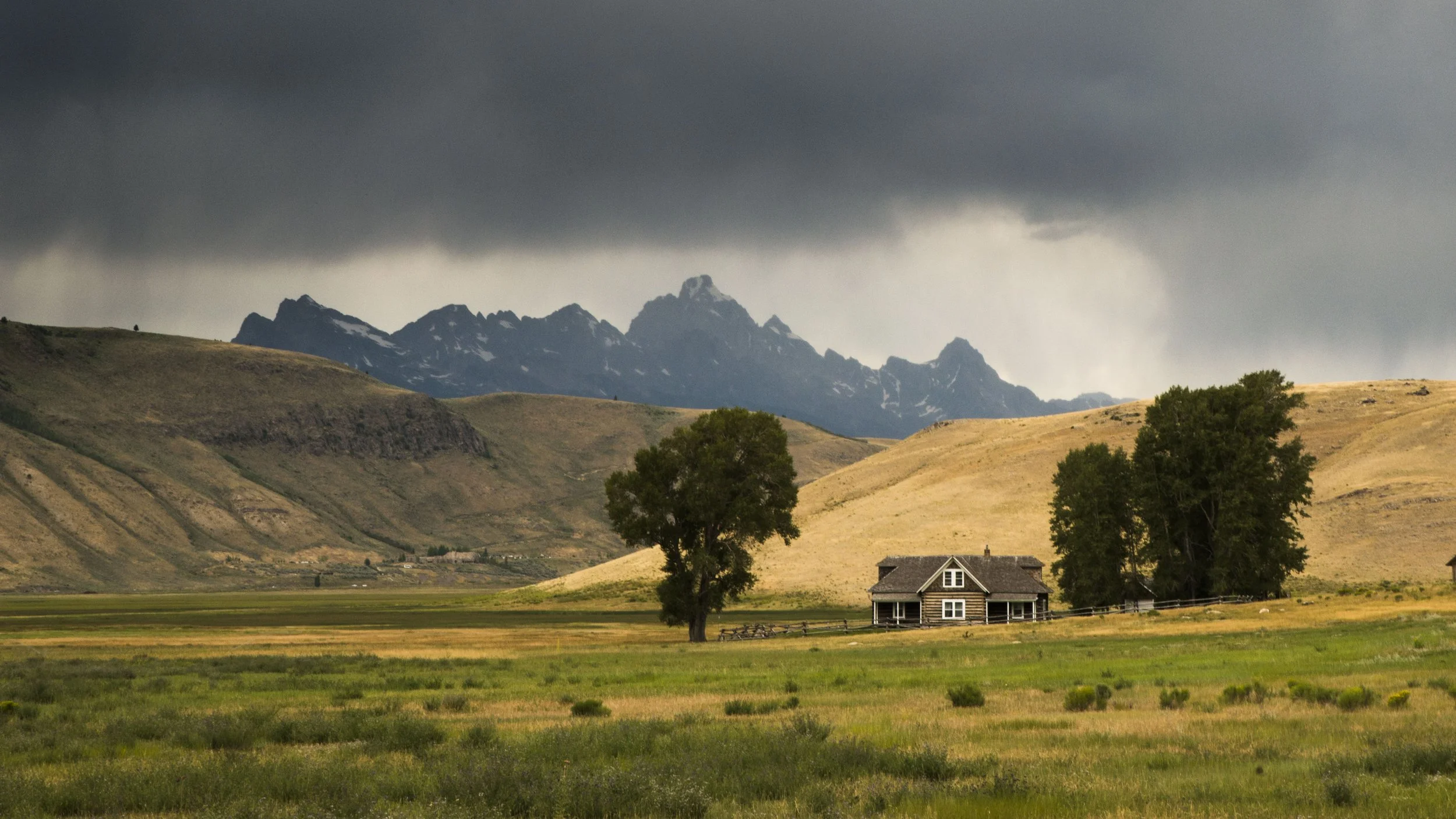 Rural landscape with a small house, rolling hills, and mountains under a cloudy sky.