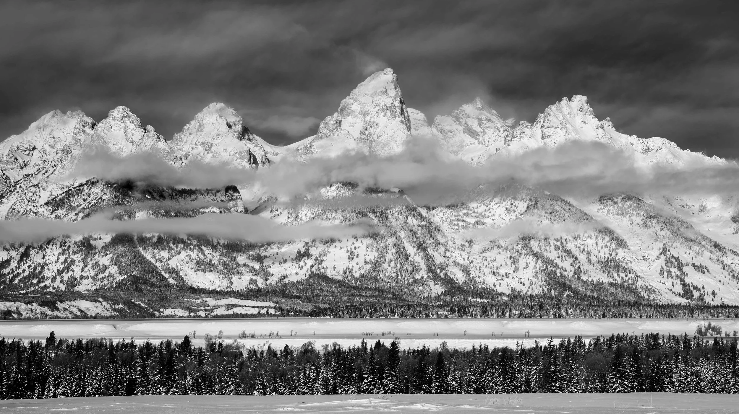 Black and white image of snow-covered mountains with clouds partially covering the peaks, dense forest at the base, and a snowfield in the foreground.