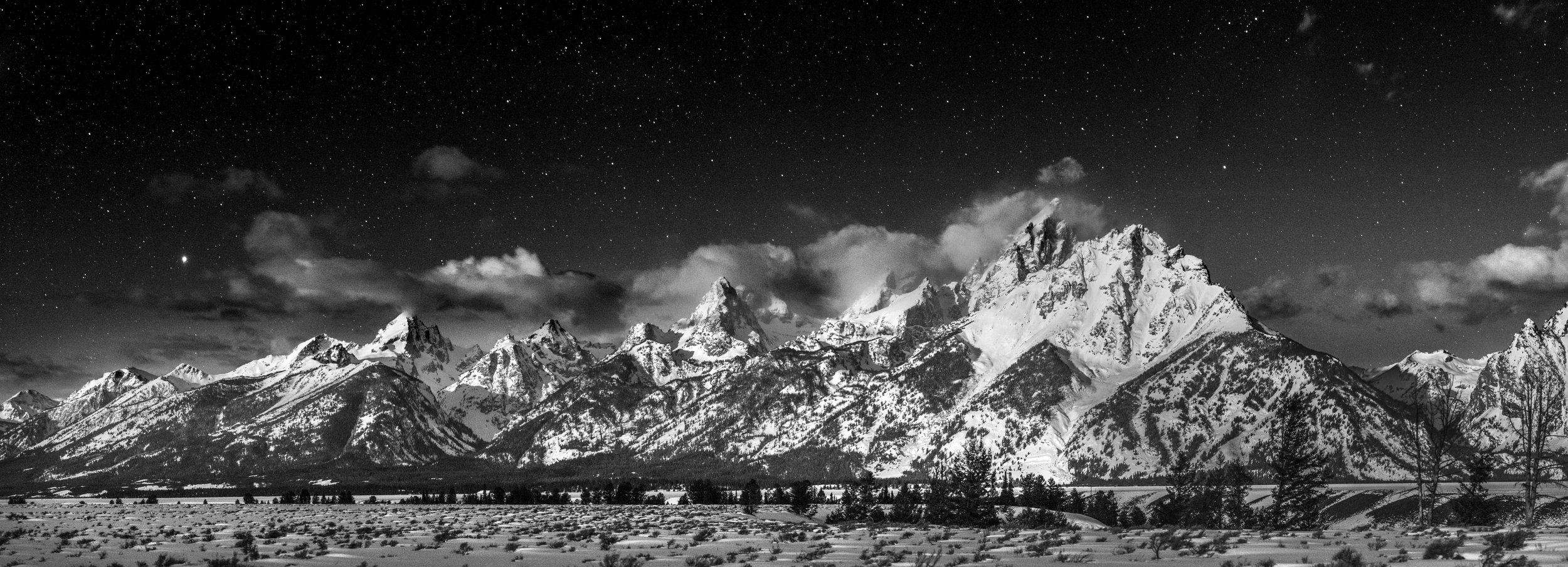 Black and white photo of snow-covered mountain range at night under a starry sky, with trees in the foreground.