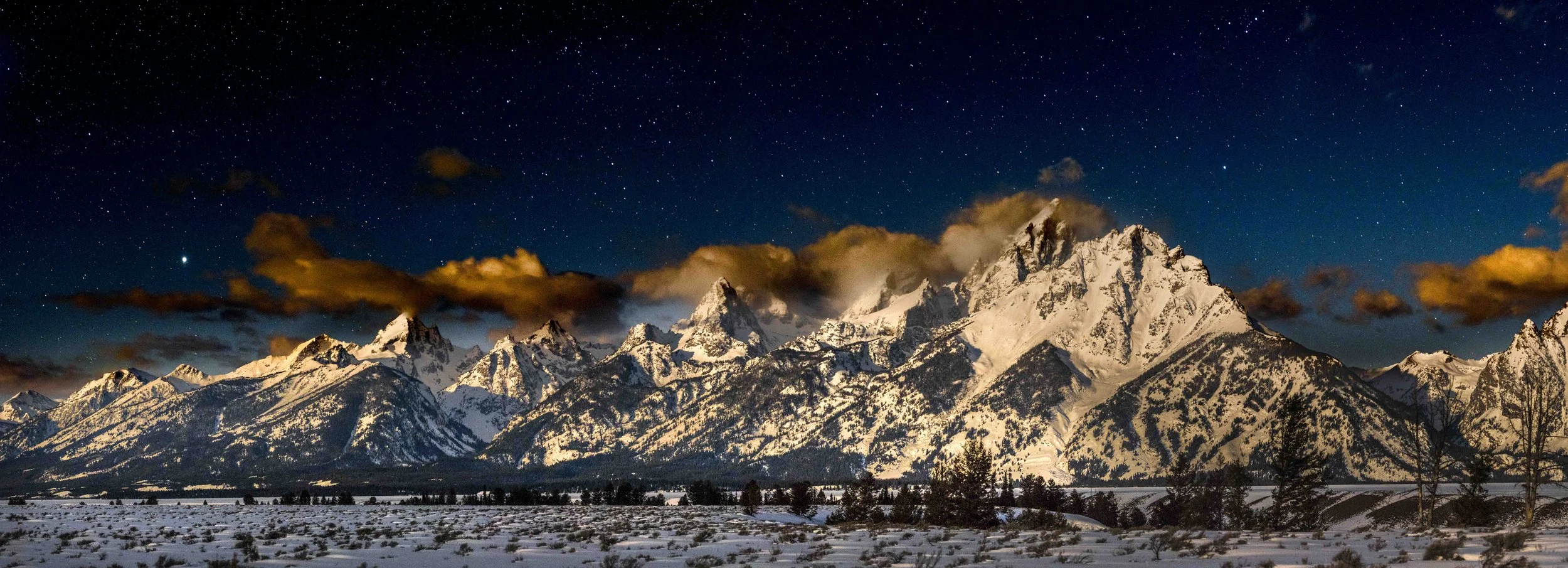 Snow-covered mountain range under a starry night sky with clouds.