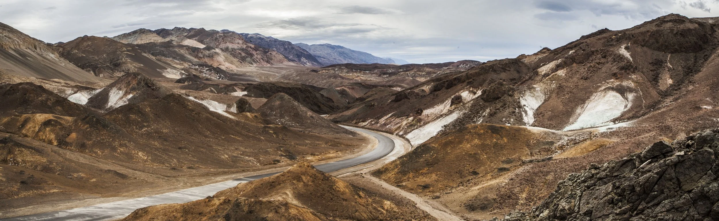 A panoramic view of a barren, mountainous desert landscape with a winding road and cloudy sky.