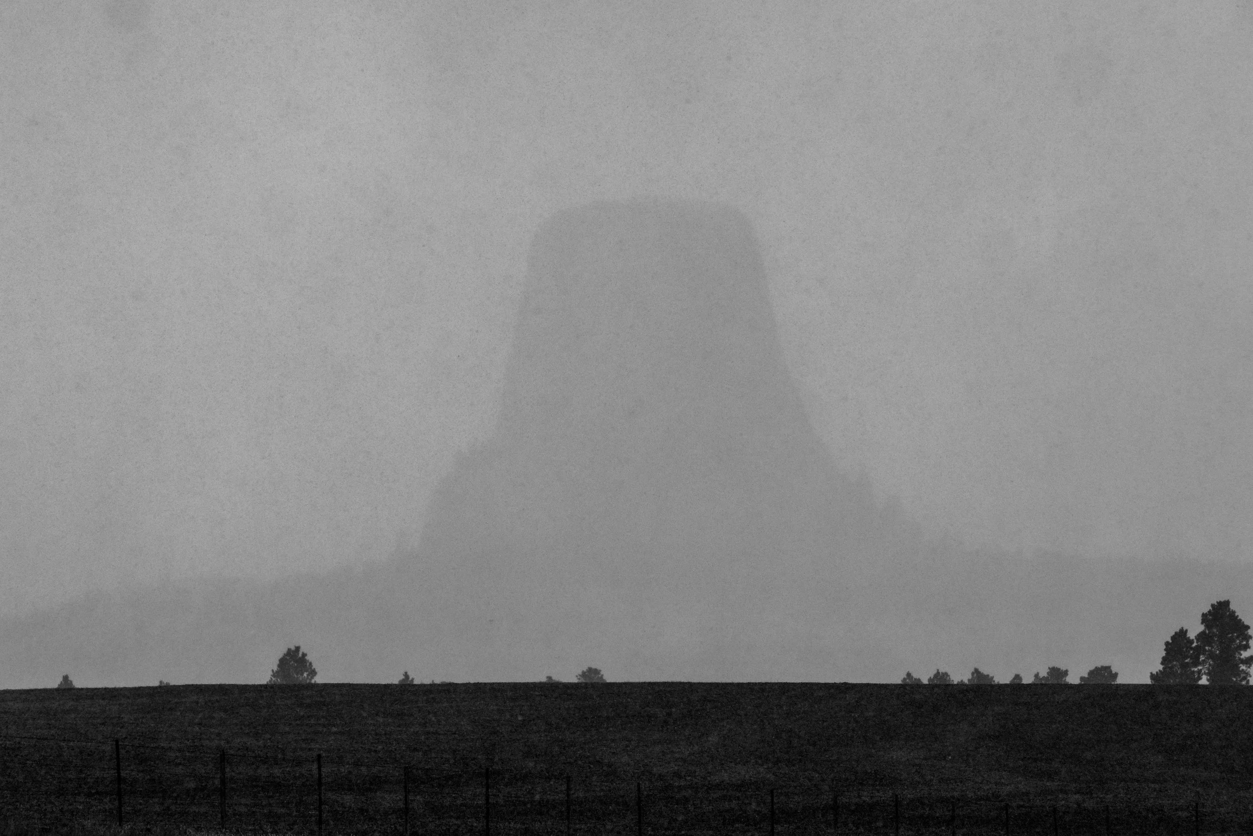 Silhouette of a large rock formation in foggy or misty conditions, with a faint horizon line and sparse trees in the foreground.