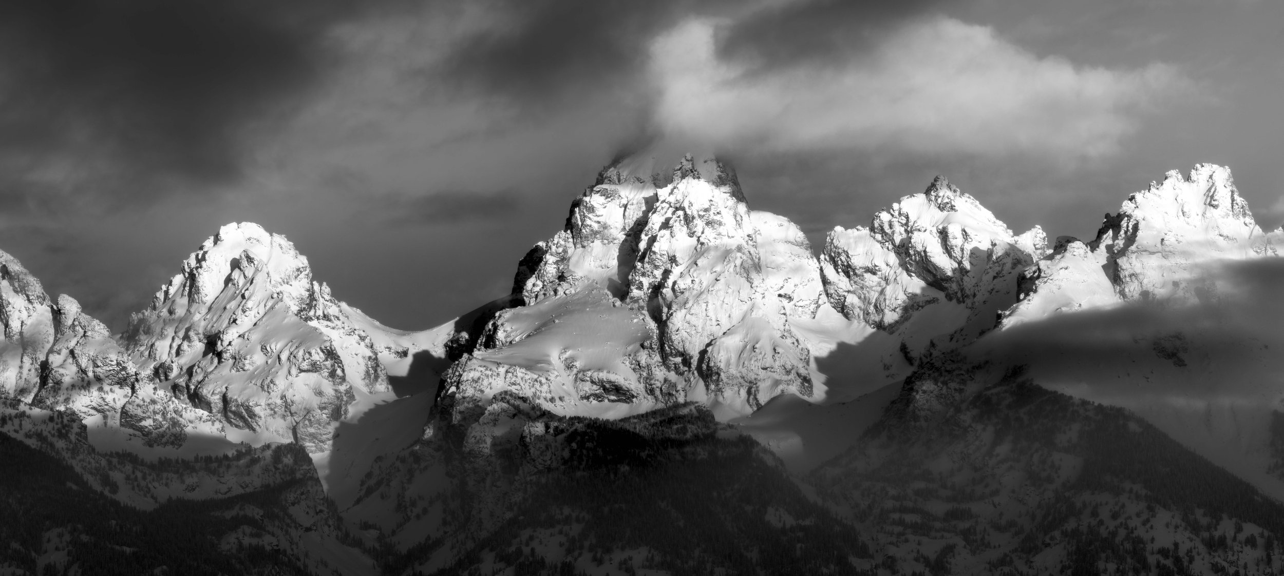 Black and white photo of snow-covered mountain peaks with dramatic clouds overhead.