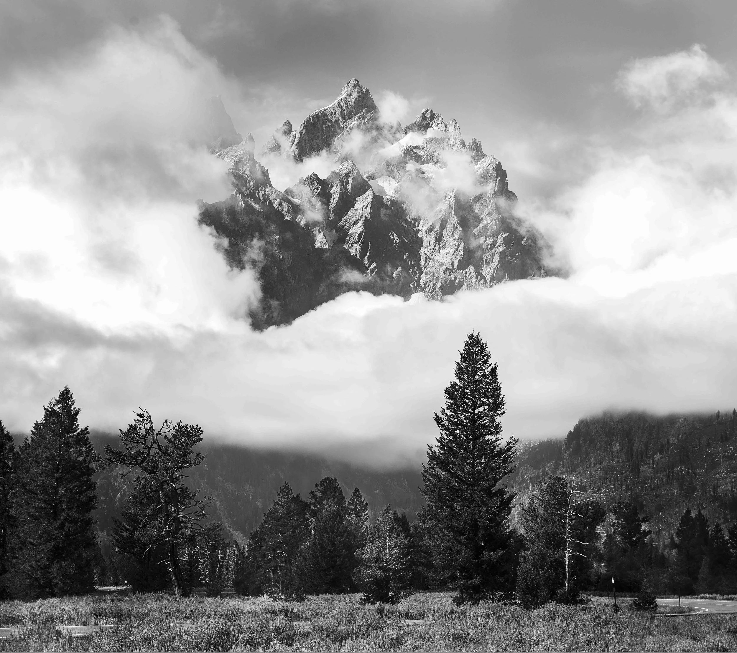Black and white photo of a mountain peak partially covered by clouds with a forested landscape in the foreground.