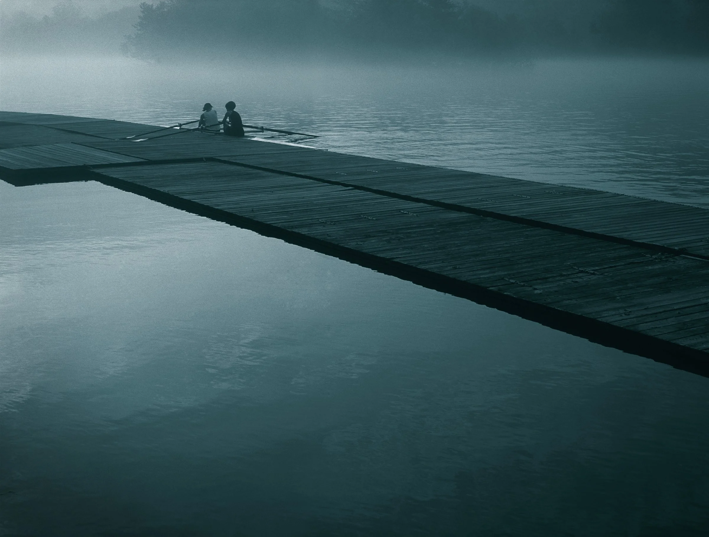 Two people rowing on a lake near a wooden dock, with misty water and trees in the background.