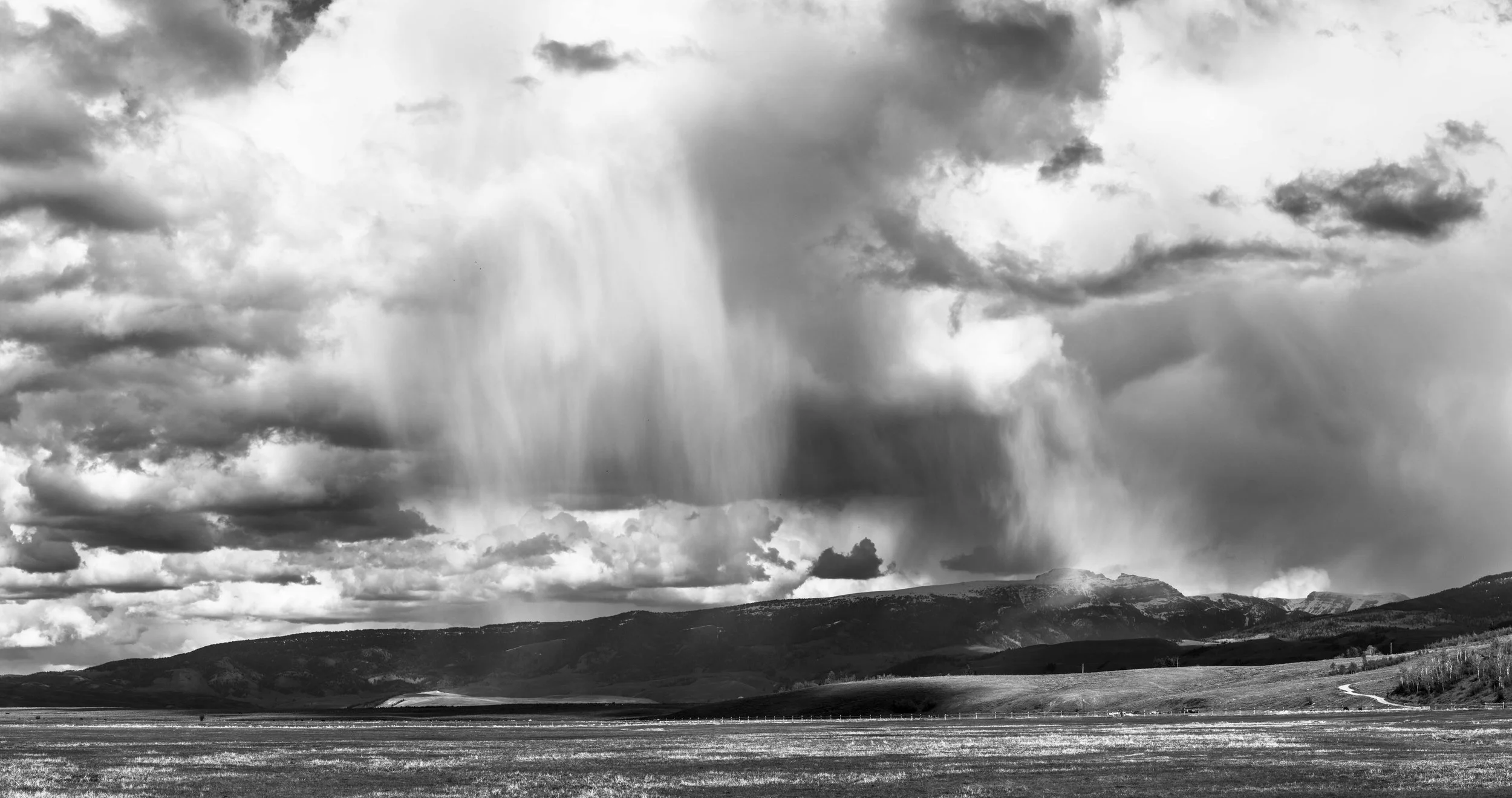 Black and white landscape with rain clouds over a mountain range and open field.