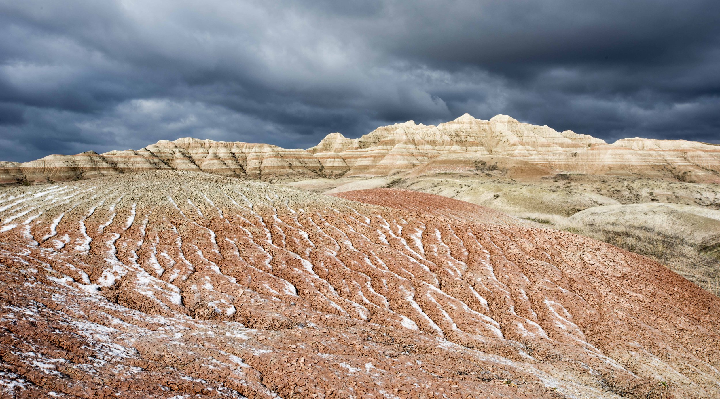 Eroded sedimentary hills under a cloudy sky, showcasing layers of red, brown, and white in a barren landscape.