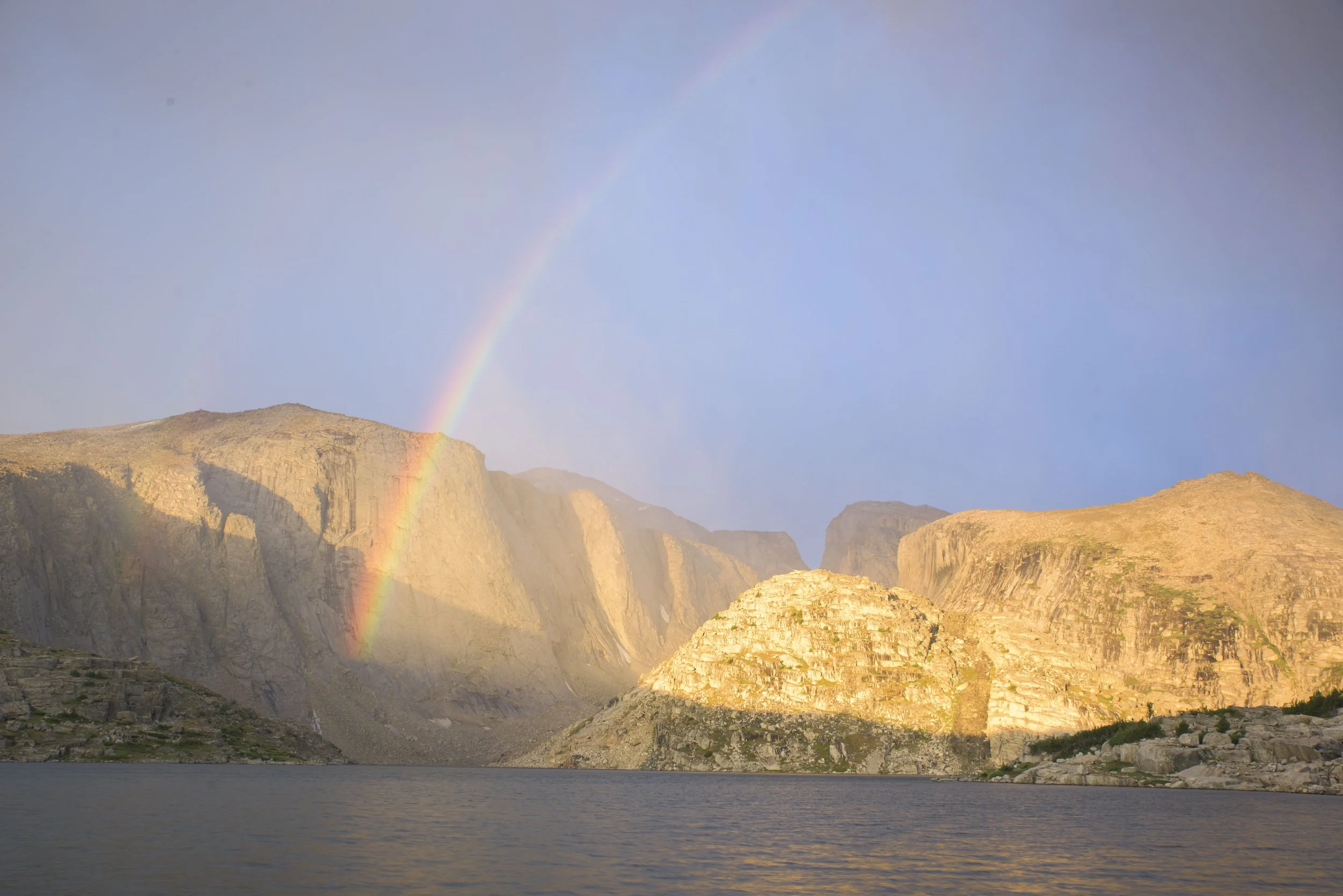 Mountain landscape with a rainbow over rocky cliffs and a lake in the foreground.