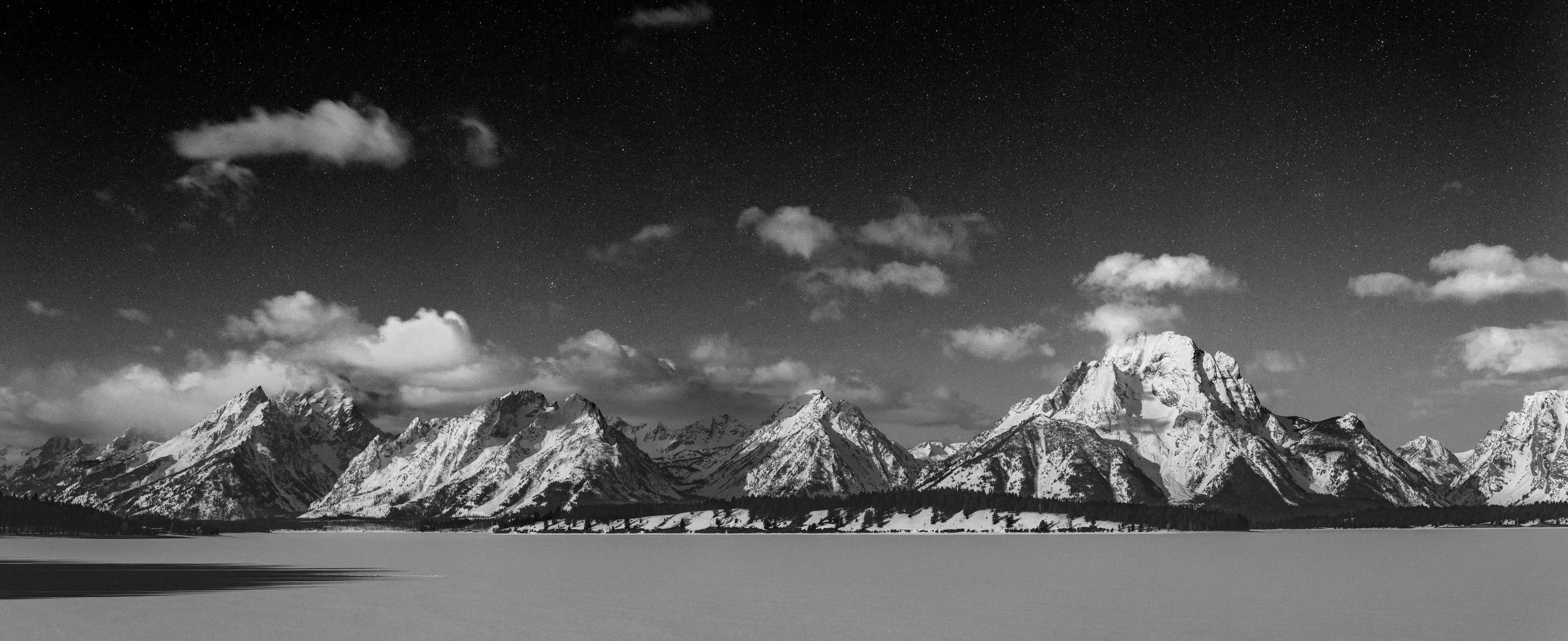 Black and white photo of snow-covered mountain range under a starry sky with scattered clouds, and a snow-covered landscape in the foreground.
