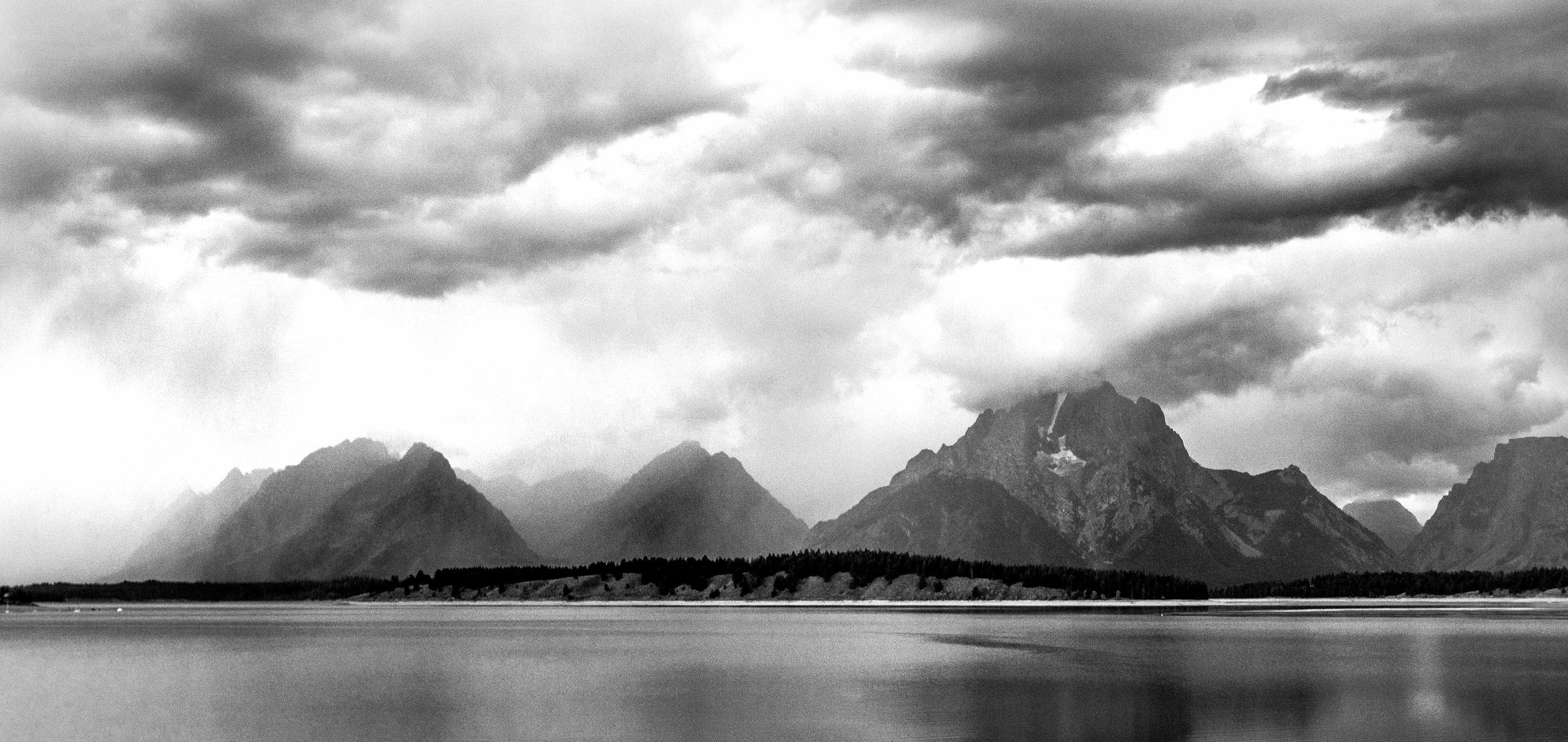 Black and white photo of majestic mountain range reflected on a calm lake, with dramatic cloudy sky.
