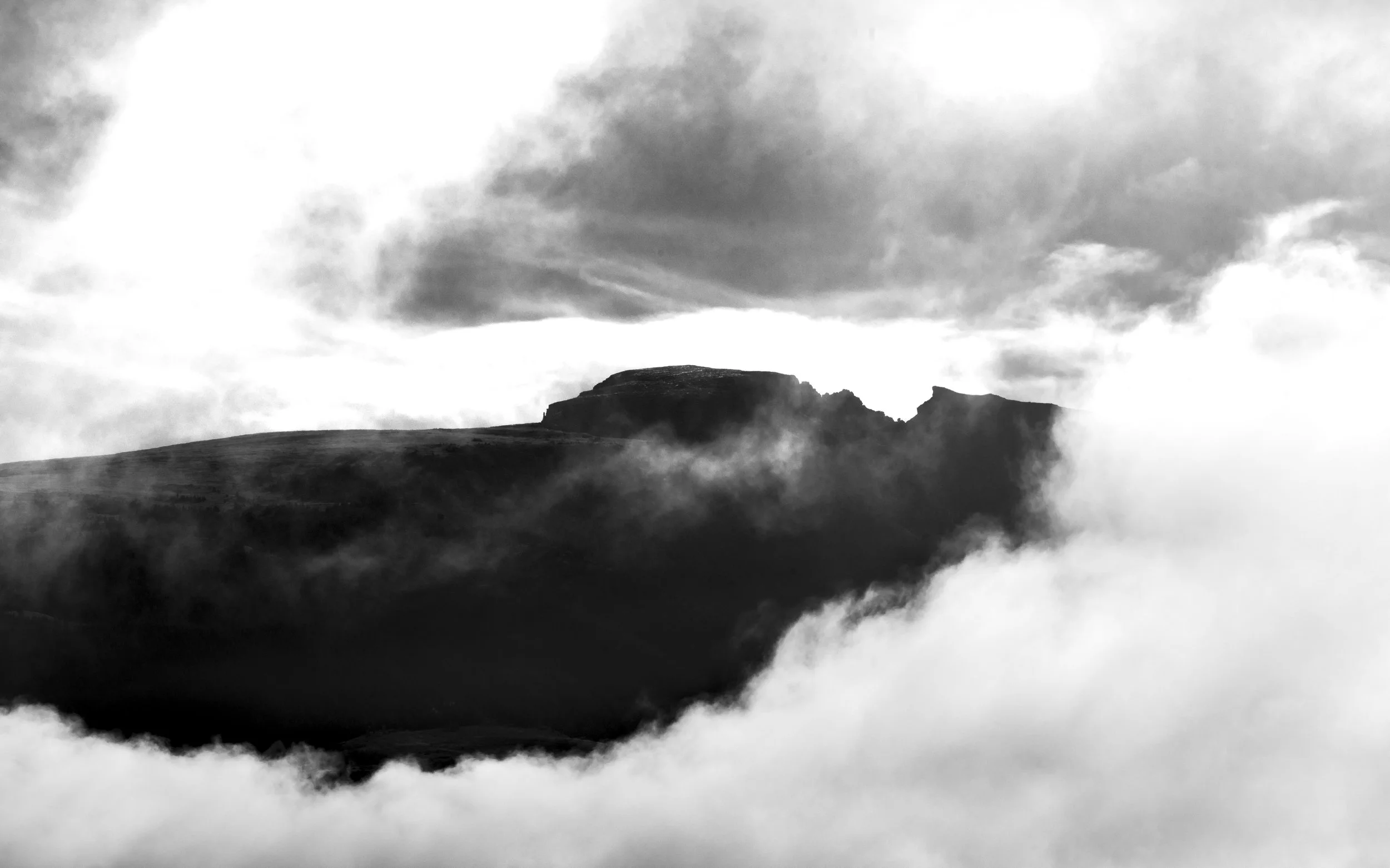 Black and white landscape photo of a mountain peak surrounded by clouds and mist under a cloudy sky.