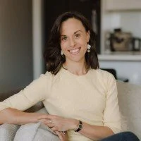 A smiling woman with dark hair wearing a cream-colored sweater, sitting on a gray sofa in a modern home interior.