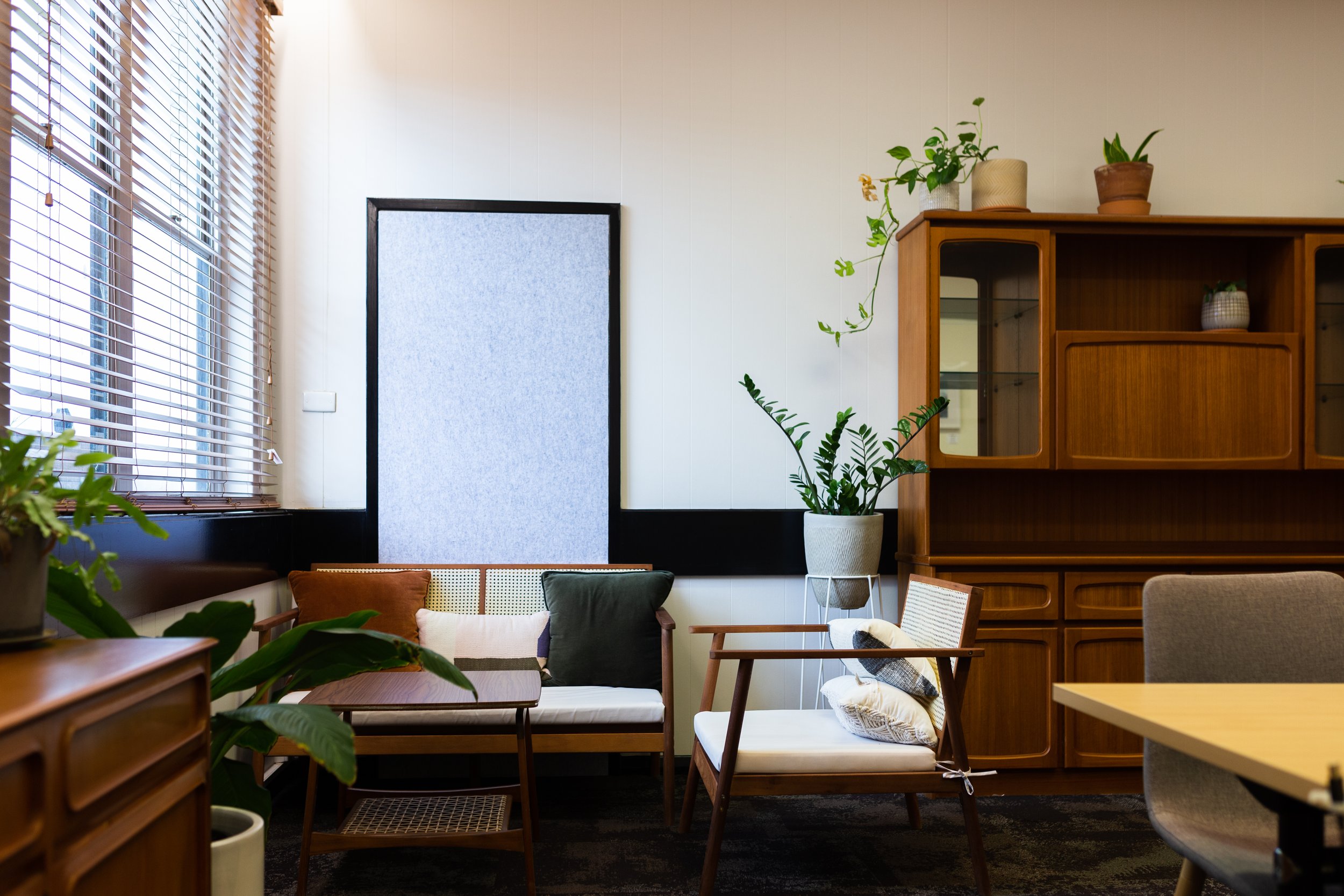 Cozy living room with wooden furniture, green plants, and natural light from windows with blinds.