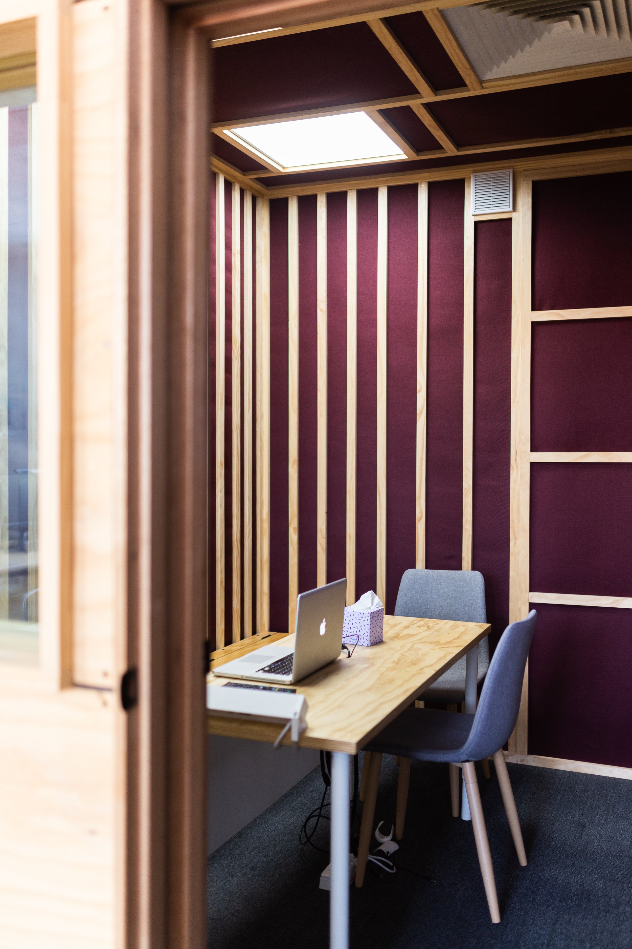 A small, private office with wooden framing and burgundy acoustic panels, containing a wooden desk with a silver laptop, a tissue box, and two gray chairs.