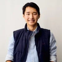 Young man smiling, wearing a blue vest over a light blue button-up shirt, standing against a plain background.